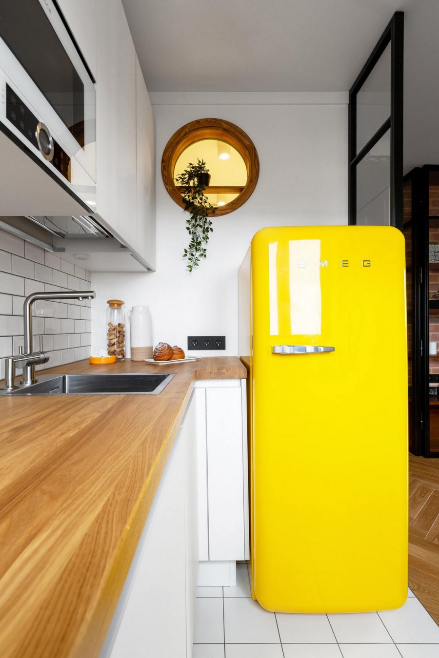 Điểm nhấn nội thất nổi bật trong phòng bếp có thể kể đến chính là chiếc tủ lạnh màu vàng tươi đến từ thương hiệu SMEG nổi tiếng. Phía trên là khung cửa nhỏ hình tròn với chậu cây xanh trang trí mềm mại.