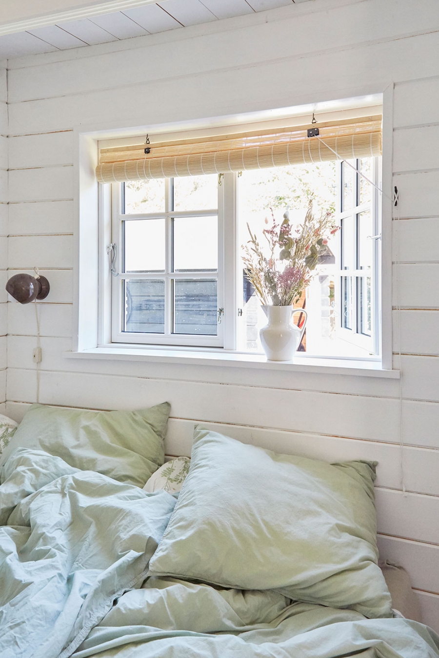 Phòng ngủ của bố mẹ với gam màu trắng chủ đạo kết hợp bộ chăn ga gối màu xanh bạc hà dịu mắt. Ô cửa sổ tích hợp mành tre để cản sáng và tạo sự riêng tư. 