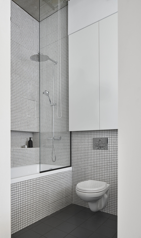 Thiết kế toilet gắn tường giúp tiết kiệm diện tích, bên trên là tủ lưu trữ kịch trần giúp tối ưu hóa không gian nhỏ. Bồn tắm nằm và tường được ốp gạch ô vuông trẻ trung, phân vùng với toilet bằng cửa kính trong suốt.
