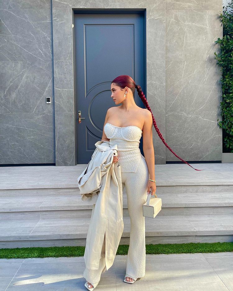 Kylie Jenner phối set quần màu trắng dệt kim với áo cúp ngực và áo khoác dài cùng tông. Điểm nhấn là mái tóc nhuộm đỏ thắt bím dài đến chấm lưng.