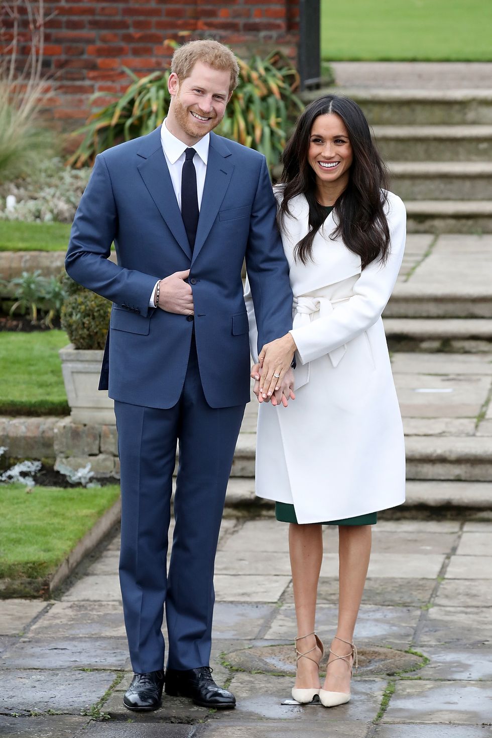 Meghan và Harry nắm tay nhau trong một cuộc gọi điện chính thức để thông báo về việc đính hôn của họ tại Sunken Gardens ở Cung điện Kensington. Cô mặc áo khoác dài màu trắng và giầy tông xuyệt tông