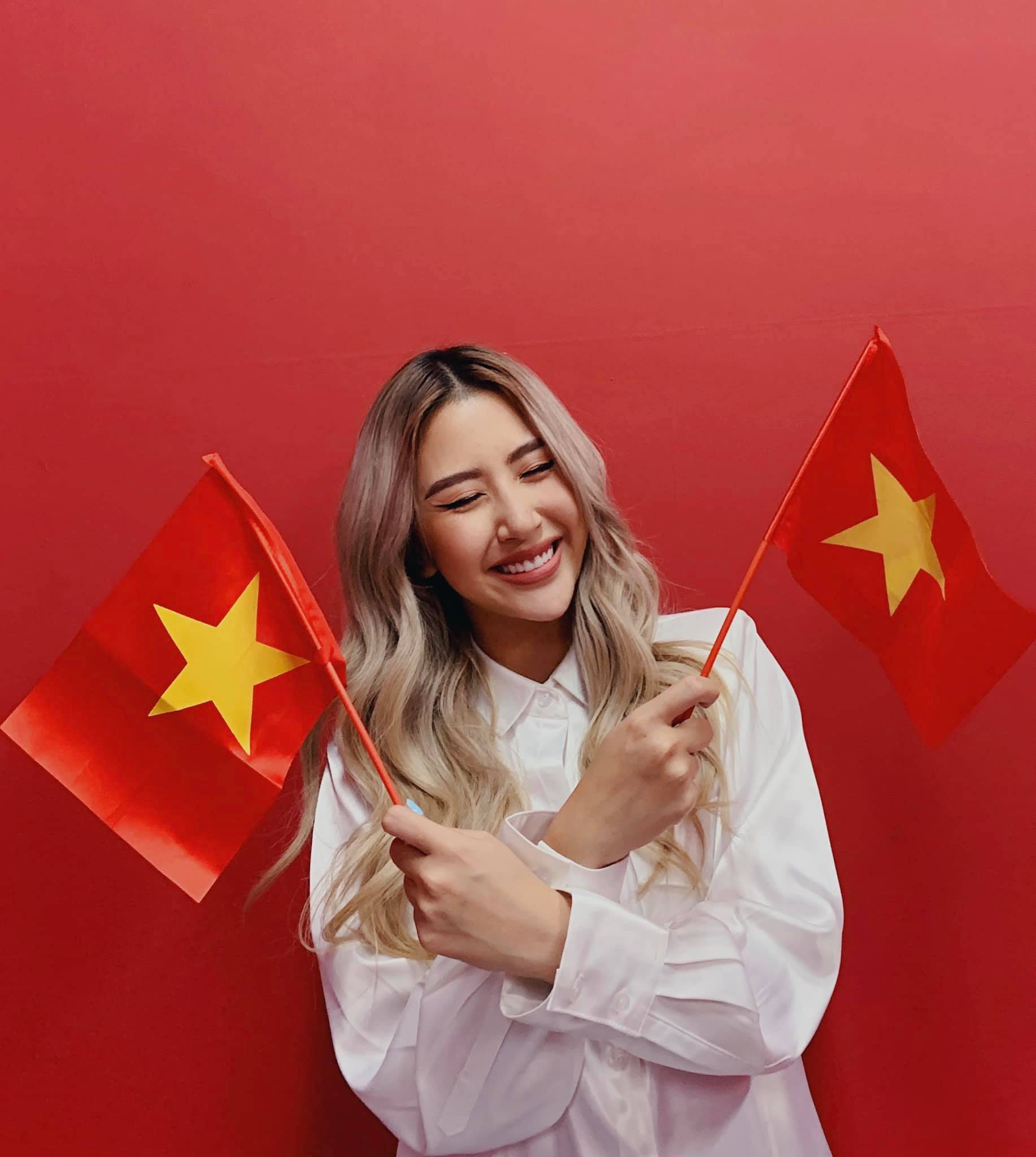 Quỳnh Anh Shyn rạng rỡ tạo dáng với background đỏ, tay cầm hai lá quốc kỳ, vui mừng đón lễ Quốc khánh bên gia đình.