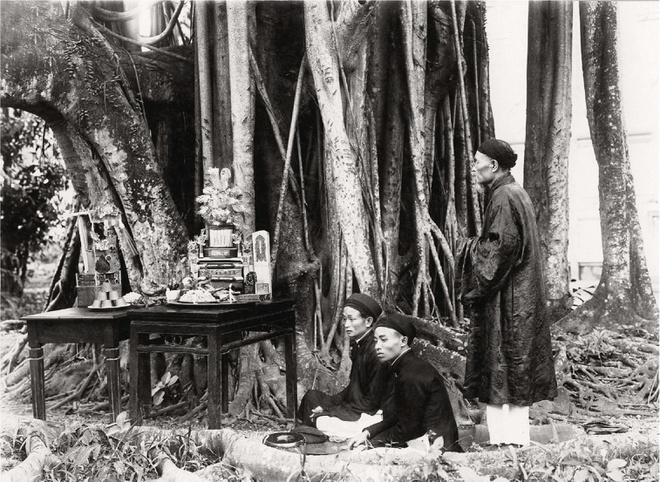 Thầy cúng đang đứng sau hai người đàn ông ngồi gọi hồn trước cây đa.