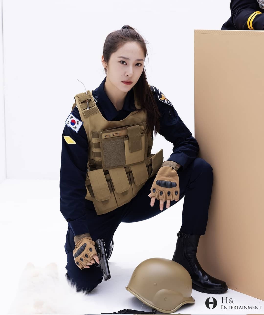 Cô cảnh sát trong bộ đồng phục cảnh vệ xanh navy, chân đi combat boot, mặc áo bảo hộ, tay cầm súng 'chất lừ'
