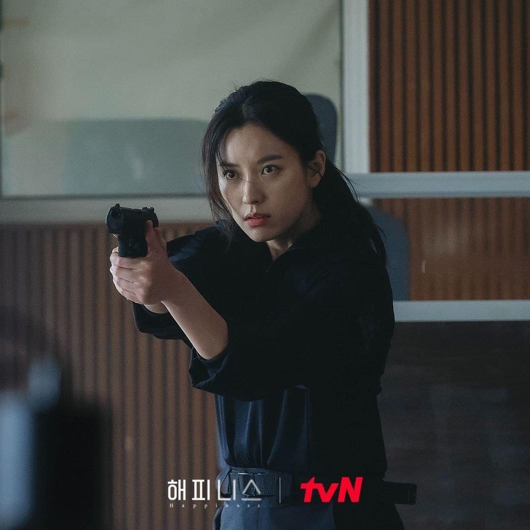 Han Hyo Joo diện set đồ đen, tóc buộc thấp đơn giản và có động tác cầm súng chĩa về phía trước đầy dứt khoát