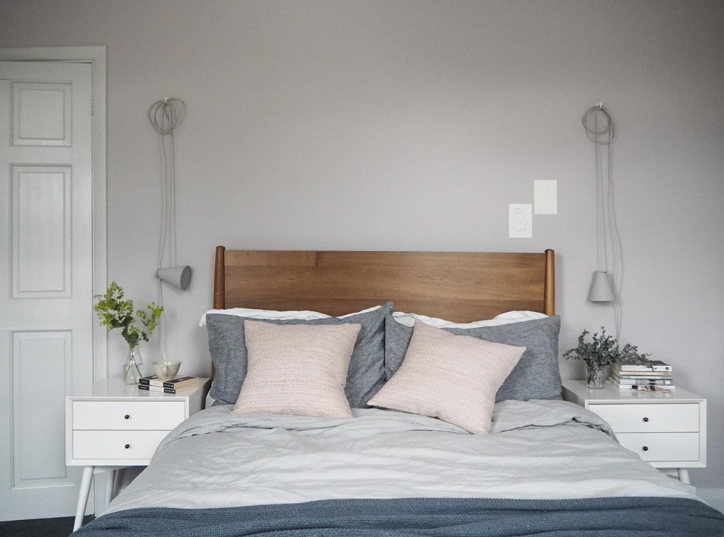 Chiếc giường ngủ là sự kết hợp giữa phong cách Mid-century đơn giản với tính hiện đại của phong cách Scandinavian. Phần đầu giường bằng gỗ đậm màu, nổi bật giữa bộ chăn ga gối nệm màu trắng, xám và hồng phớt dịu nhẹ.