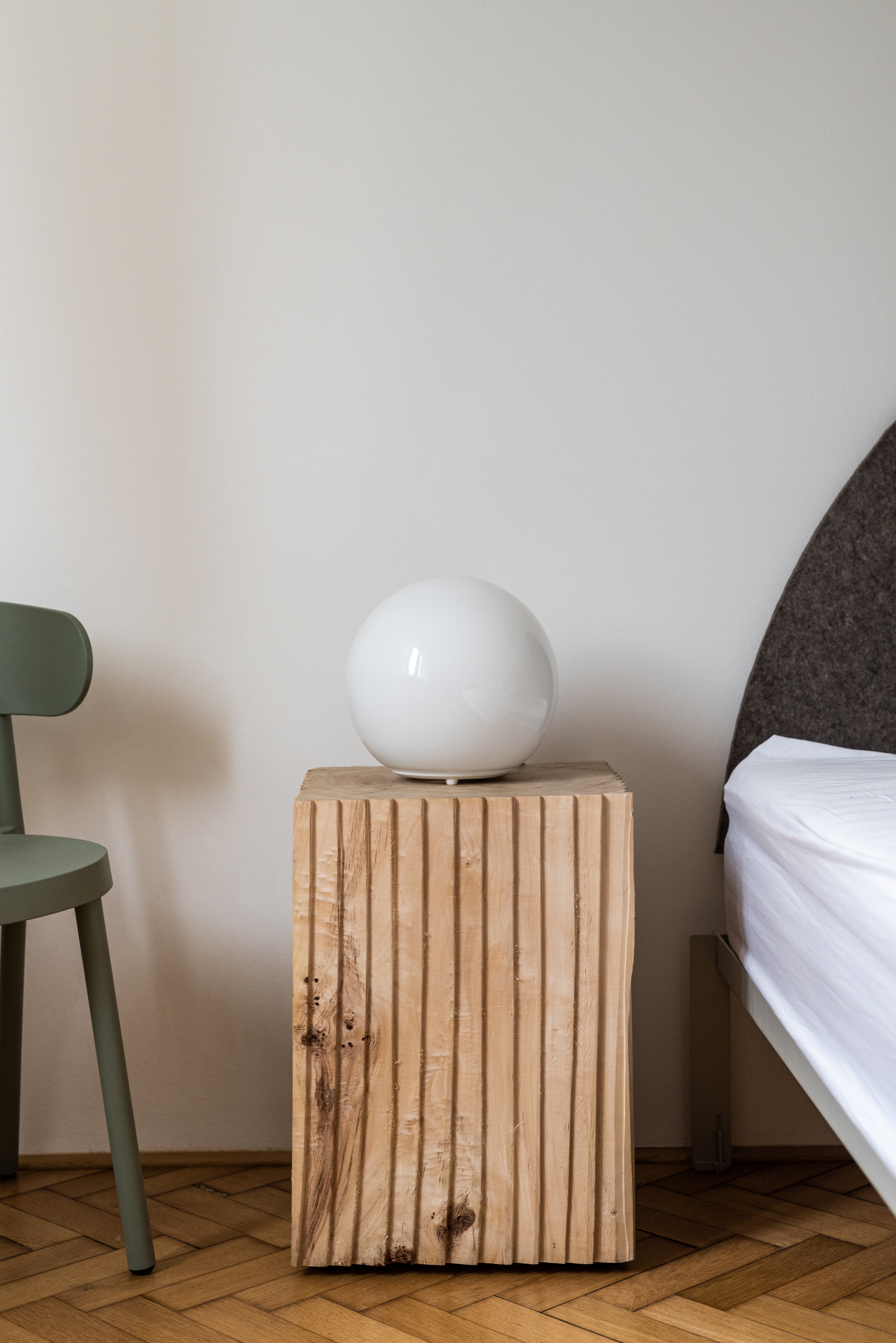 Hai khối gỗ tự nhiên đặt đối xứng nhau thay cho chiếc táp đầu giường quen thuộc. Bên cạnh là chiếc ghế màu xanh làm điểm nhấn cho căn phòng ngập tràn sắc trắng.
