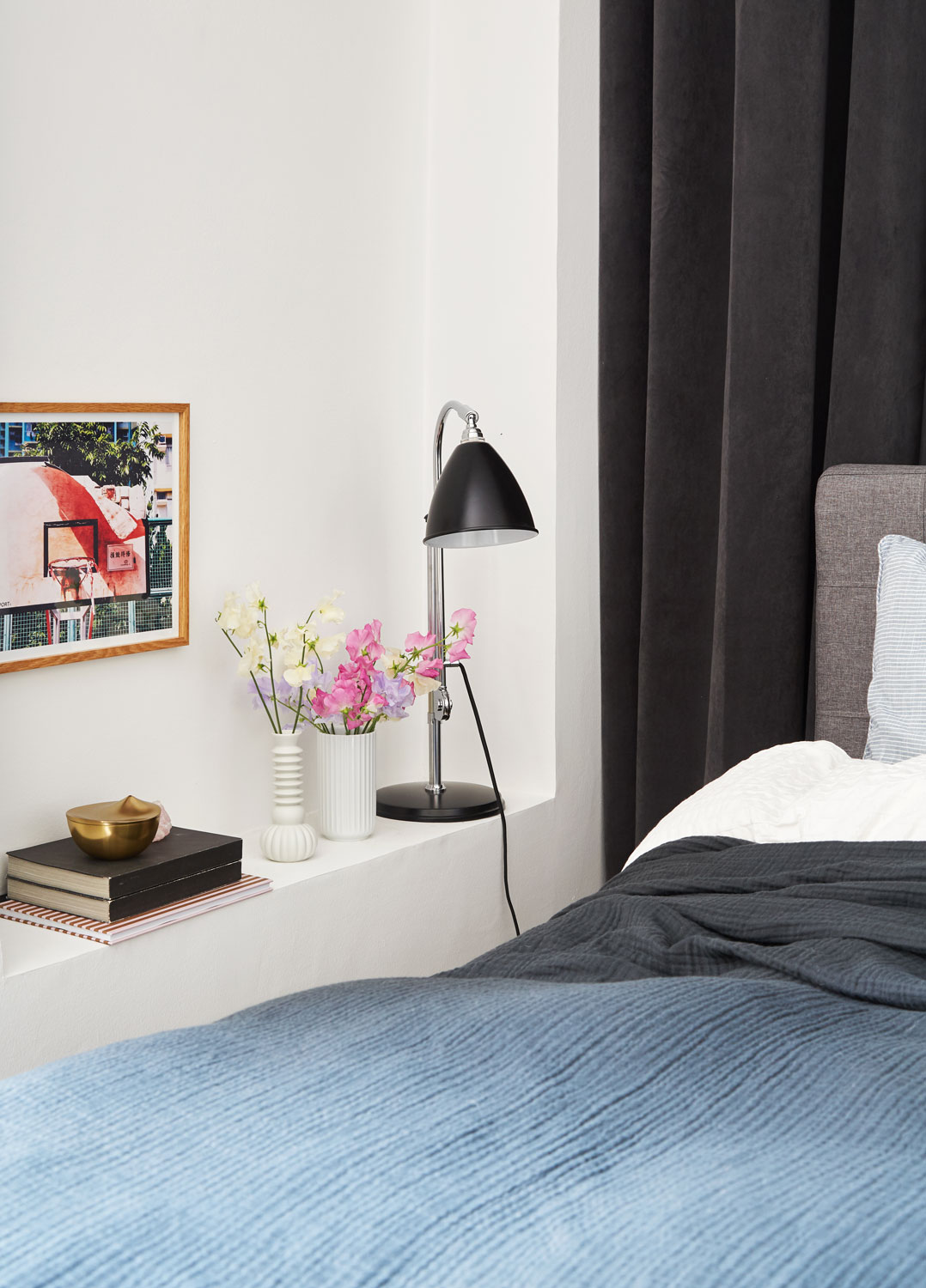 Mảng tường bên trái giường ngủ được tận dụng phần hốc để sắp xếp đèn ngủ có thiết kế màu đen, dáng thanh mảnh và hiện đại, hai lọ hoa sứ xinh xinh, một ít sách và bức tranh trang trí cho nền tường không còn buồn tẻ. Bộ chăn ga gối với màu chủ đạo màu xanh lam đậm và xám - một lựa chọn tuyệt vời để tạo ra cảm giác bình yên. Riêng tấm chăn mỏng được phủ che kín tận chân giường mang lại vẻ nhẹ nhàng và như được che chở.