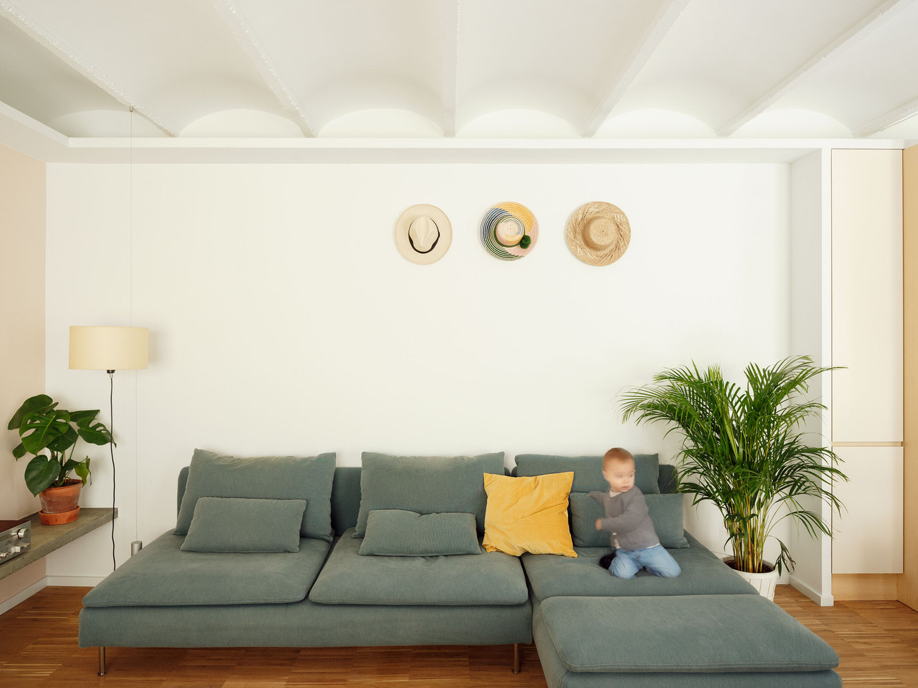 Phòng khách được bố trí chiếc ghế sofa chữ L màu xám không tay vịn - xu hướng thiết kế nội thất được ưa chuộng vì tạo cảm giác rộng rãi cho không gian. Những chiếc mũ nhiều kiểu dáng cũng giúp cho cho bức tường phòng khách sinh động hơn.