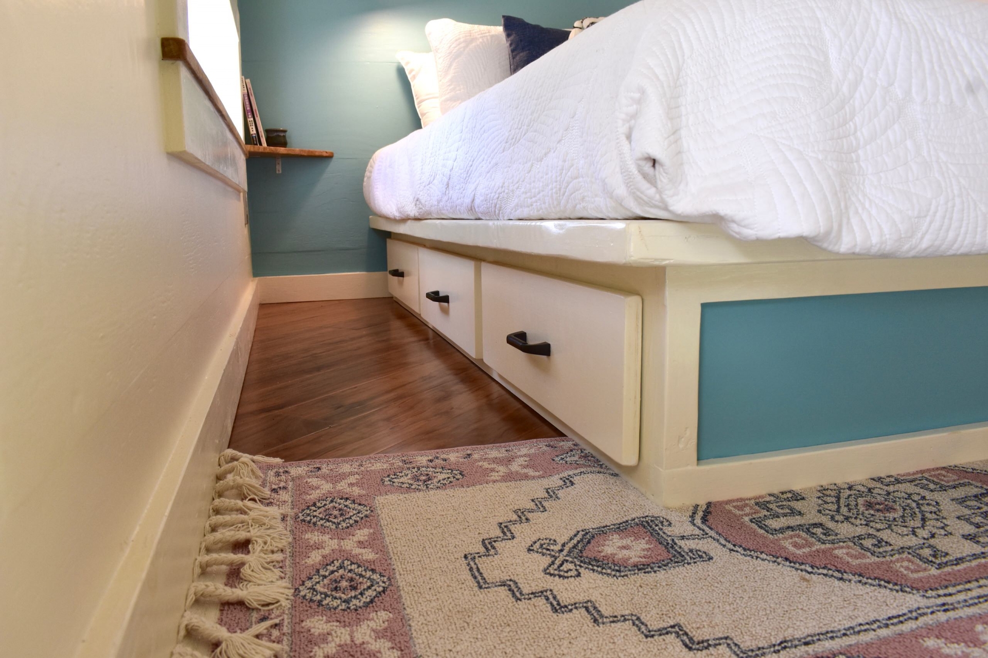 Chiếc giường tích hợp các hộc lưu trữ ẩn bên dưới để tăng khả năng lưu trữ đồ đạc cho không gian luôn gọn gàng. Sàn gỗ ấm áp với tấm thảm dệt họa tiết thổ cẩm nhẹ nhàng, vừa giữ vệ sinh vừa làm điểm nhấn cho phòng ngủ.