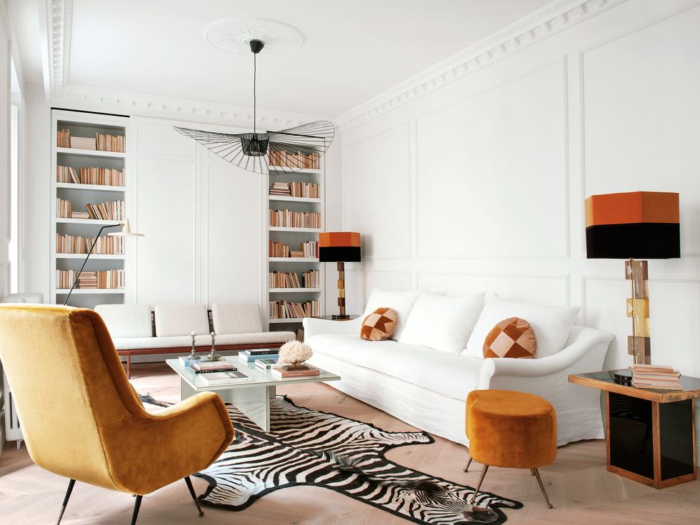 Phòng khách xinh đẹp với bộ sofa màu trắng êm ái, nổi bật chiếc ghế bành và ghế đôn màu cam gạch. Tấm thảm trải sàn họa tiết da ngựa vằn độc đáo kết hợp với chiếc đèn trần với thiết kế mềm mại duyên dáng.