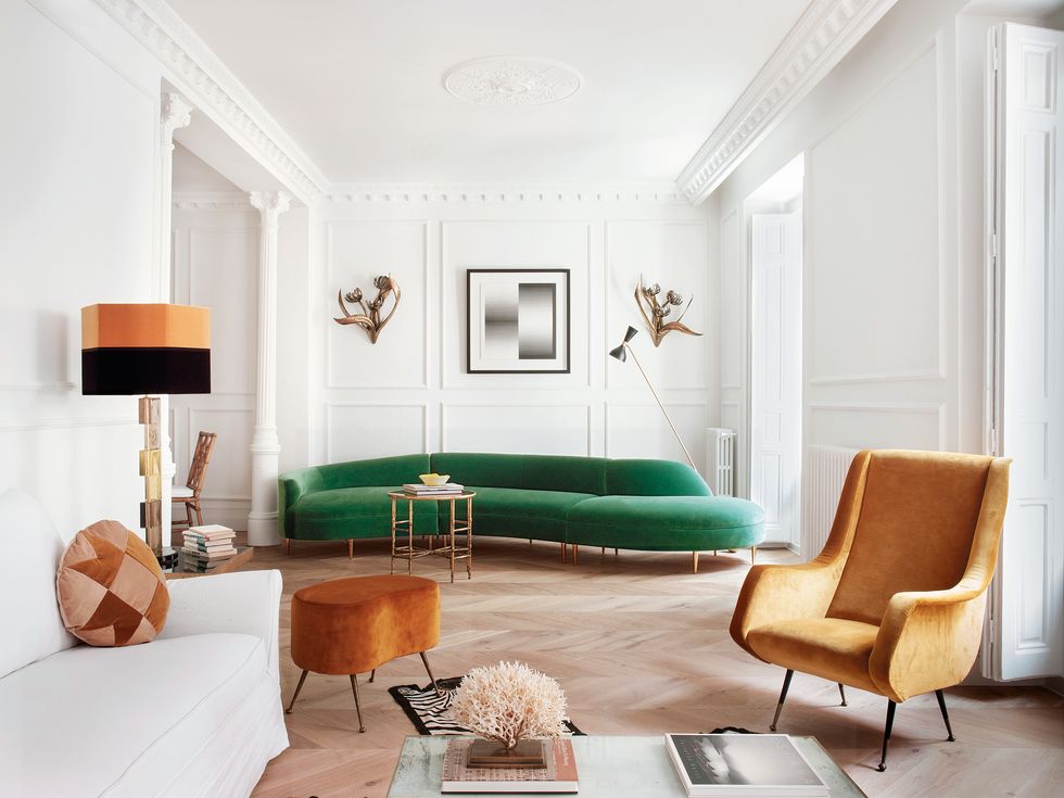 Đường nét uốn lượn của chiếc ghế sofa màu xanh nổi bật với chất liệu vải nhung sang trọng. Bức tường phía sau sofa được tô điểm bằng những bông hoa bằng vàng đồng phong cách cổ điển và bức tranh của Getulio Alviani đặt ở trung tâm.