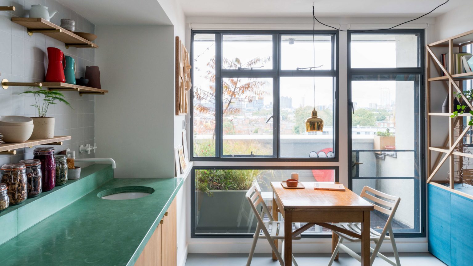 Một chiếc tủ sơn xanh lam kết hợp kệ gỗ kiểu lưới mắt cáo, đóng vai trò làm vách ngăn phòng bếp với phòng khách. Khoảng trống giữa hai khu vực này dành cho bộ bàn ăn bằng gỗ đơn giản và mộc mạc.