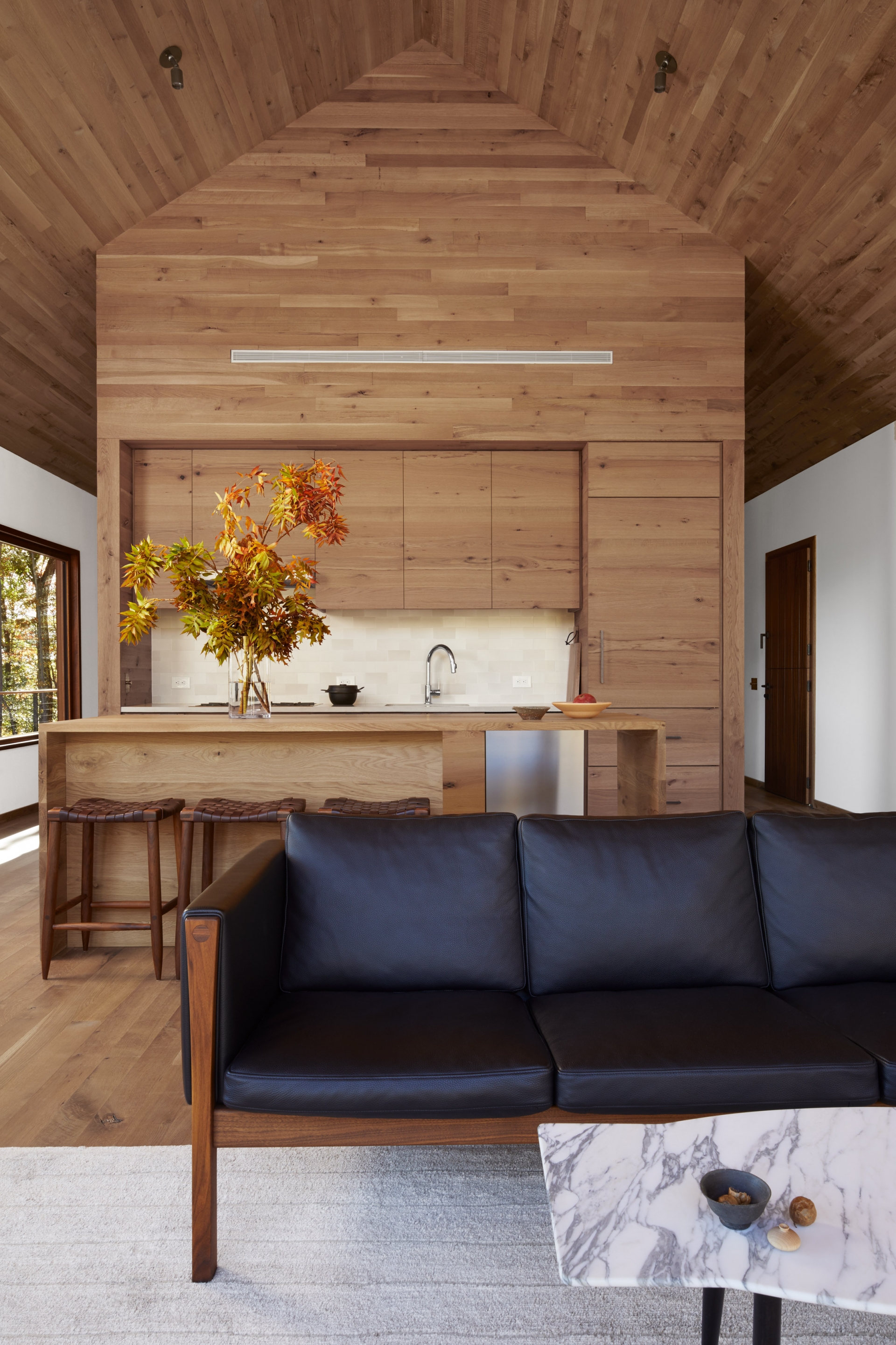 Bộ ghế sofa khung gỗ đệm da màu đen cho cảm giác vững chãi và mạnh mẽ. Phía sau là phòng bếp với hệ tủ bếp và đảo bếp bằng gỗ đẹp mắt. 