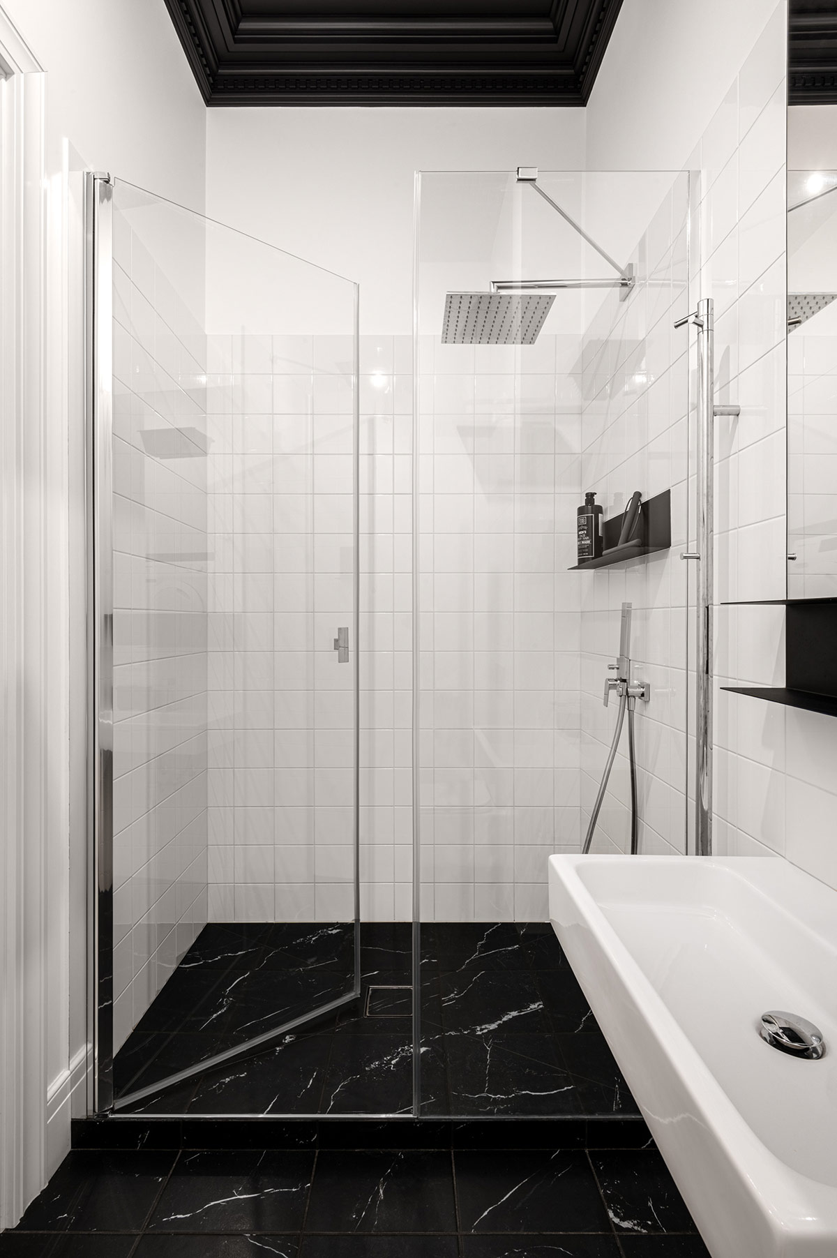 Toilet và bồn rửa tay đều được thiết kế gắn tường để giúp phòng tắm rộng rãi và dễ dàng vệ sinh sàn nhà. Buồng tắm được phân tách với phòng vệ sinh bằng cửa kính trong suốt, chiếc kệ tối giản màu đen chỉ đặt vật dụng thật cần thiết cho khu vực này thông thoáng hơn.