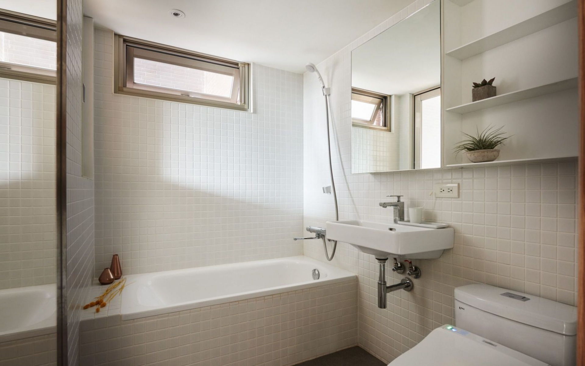 Nội thất phòng tắm vẫn “trung thành” với gam màu trắng nhưng có thêm cây xanh trang trí. Buồng tắm đứng trước khi cải tạo được thay thế bằng bồn tắm nằm. Tấm gương phản chiếu ánh sáng từ ô cửa giúp phòng tắm rộng và thoáng đãng hơn.