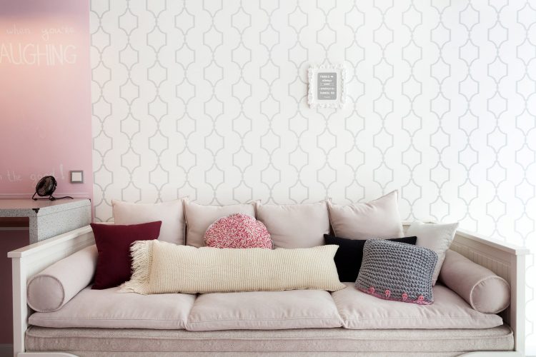 Phòng khách với bức tường trắng họa tiết hình học lạ mắt cùng chiếc ghế trường kỷ với lớp đệm dày và gối tựa đủ sắc màu êm ái.