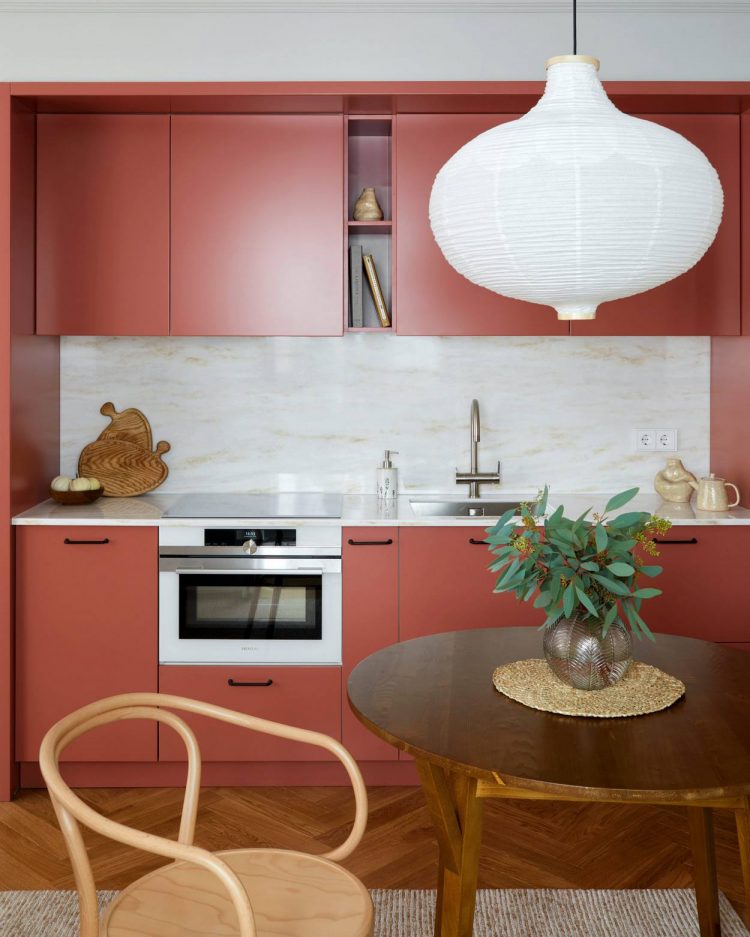 Phòng bếp sử dụng gam màu đỏ san hô chủ đạo cho cả tủ bếp trên và dưới mang đến cái nhìn cực kỳ nổi bật. Đi kèm với nó là màu đá tự nhiên của khu vực backsplash, đèn lồng treo màu trắng cũng như sắc xanh lá trên bàn ăn gỗ.