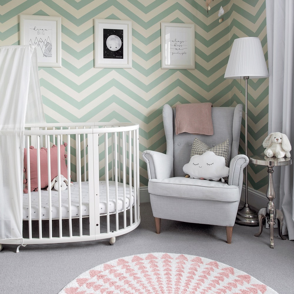 Phòng ngủ của em bé nhỏ với giấy dán tường họa tiết hình zig zag màu xanh bạc hà và màu be nhạt dễ thương. Chiếc cũi màu trắng có lắp bánh lăn tiện ích. Chiếc ghế bành màu xám để bố mẹ có thể trông chừng em bé.