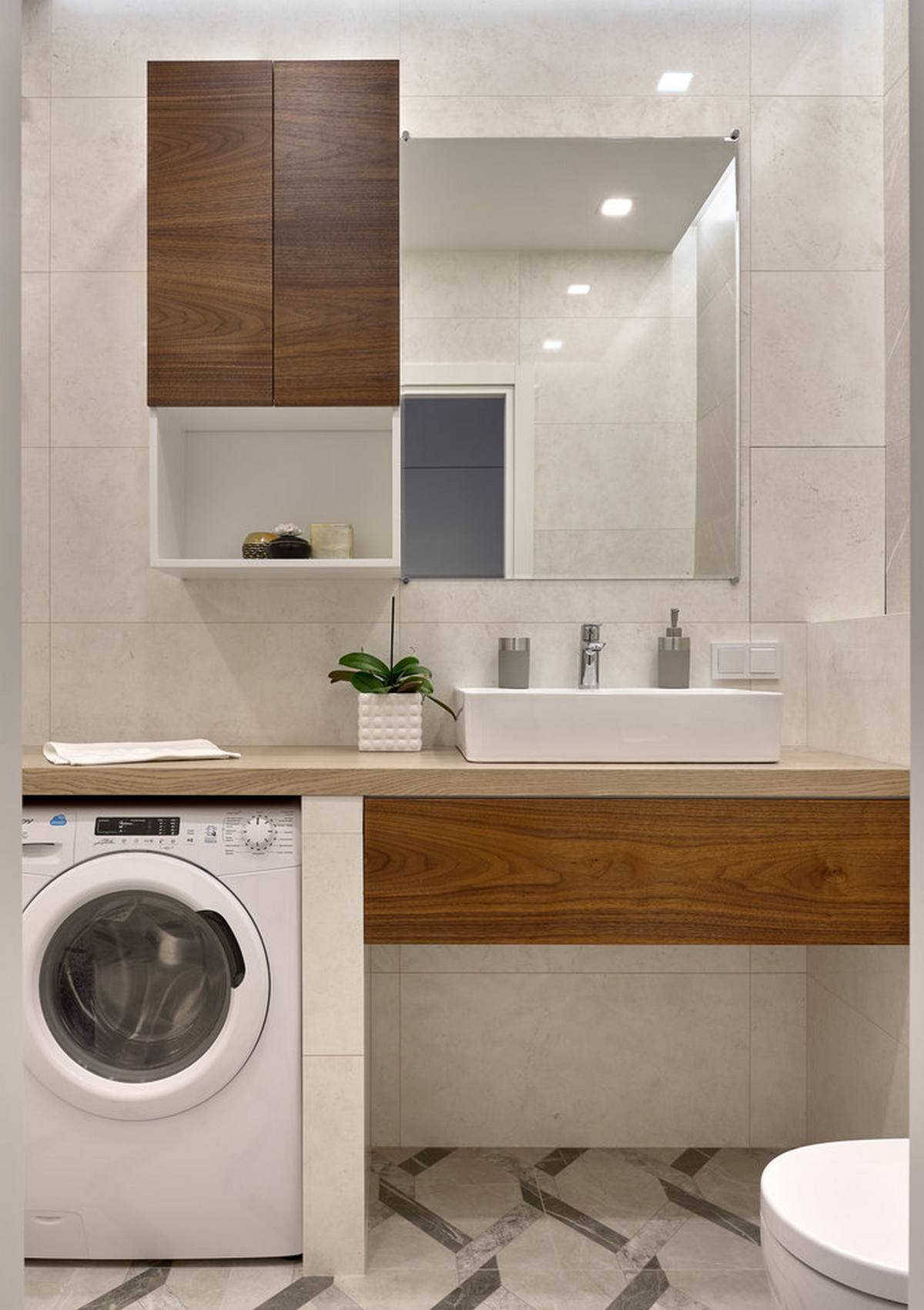 Tấm gương hình chữ nhật treo dọc bức tường giúp 'cơi nới' phòng tắm, bên dưới kệ đặt bồn rửa là khu vực dành cho chiếc máy giặt nhỏ nhắn, đáp ứng đủ yêu cầu sử dụng của hai vợ chồng.