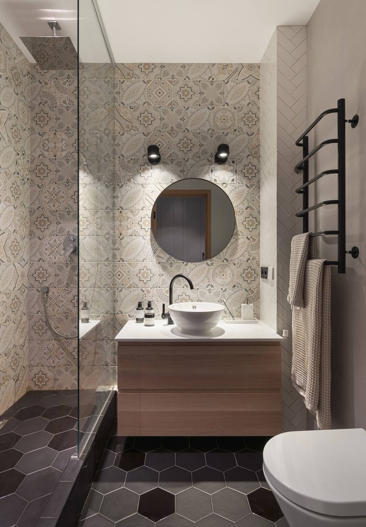 Bề mặt tường phòng tắm ốp gạch bông cổ điển. Tủ kết hợp bồn rửa hiện đại, chiếc thang treo khăn tắm gắn tường cũng như tấm kính trong suốt phân vùng buồng tắm và toilet cho căn phòng được thông thoáng bất chấp diện tích nhỏ hẹp.