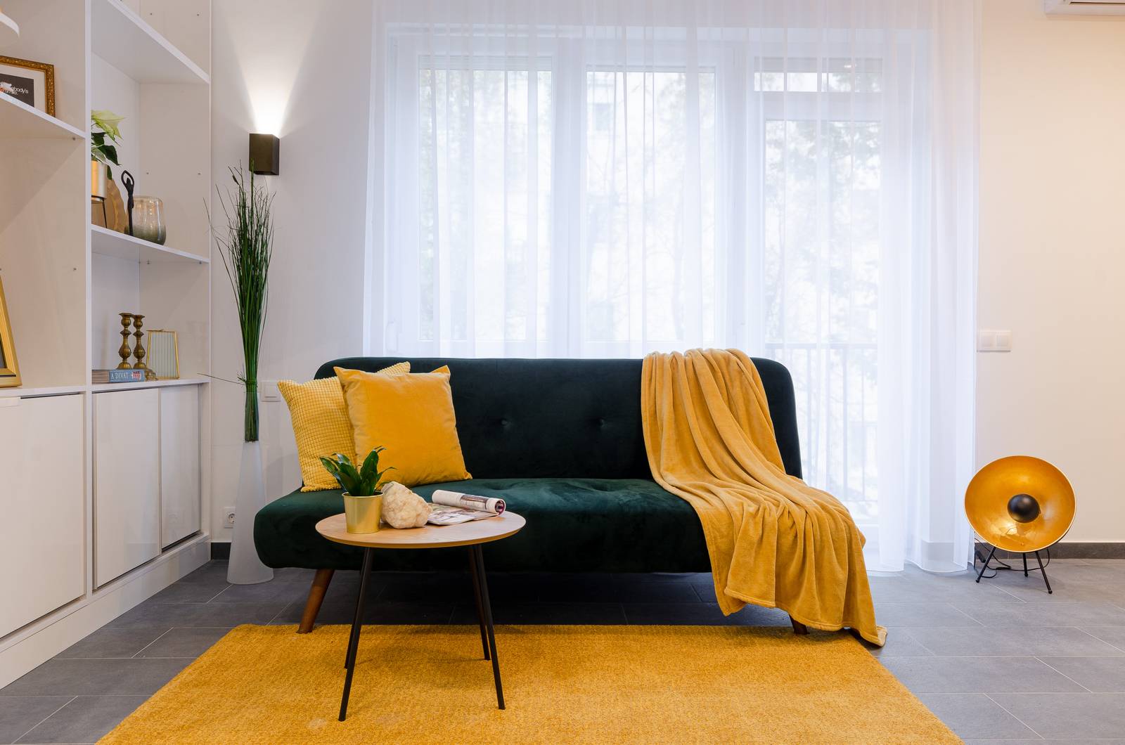 Phòng khách nổi bật với ghế sofa xanh ngọc lục bảo thời thường, khi kết hợp cùng thảm trải sàn, gối tựa và chăn mỏng màu vàng mù tạt thì càng thêm ấn tượng.