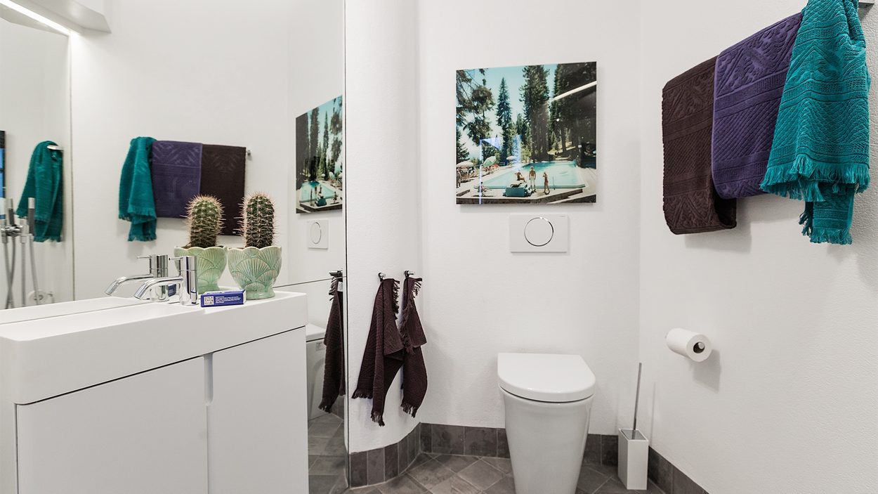 Phòng tắm sáng sủa nhờ sơn tường màu trắng, sàn lát gạch màu xám nhạt và tấm gương soi cỡ lớn ốp gần như trọn vẹn bức tường sau bồn rửa.