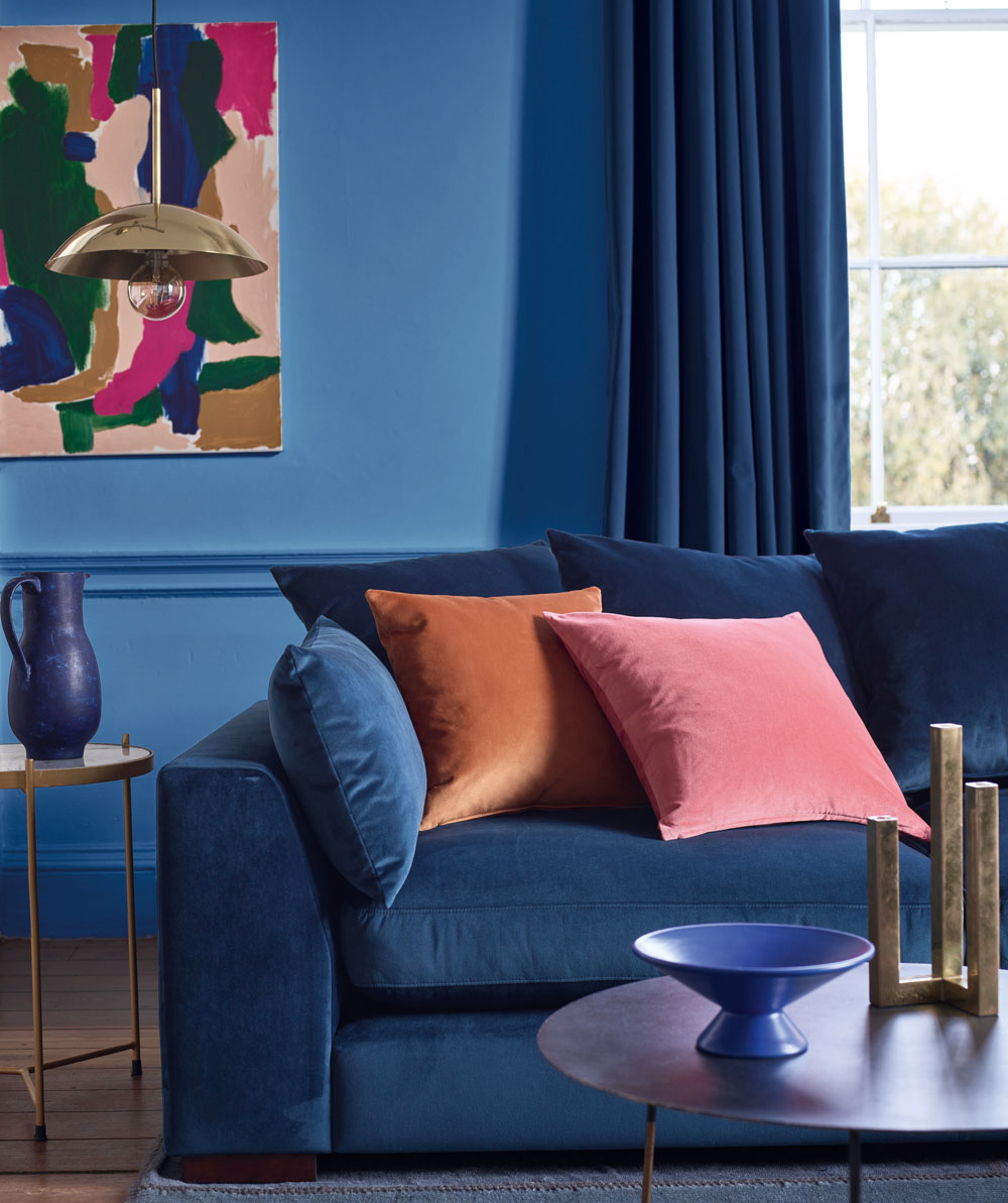 Bức tường xanh lam kết hợp với rèm cửa sổ, ghế sofa bọc nhung êm ái, thêm chút sắc hồng và cam gạch của gối tựa và tranh treo tường tăng thêm điểm nhấn ấm áp cho phòng khách trở thành một “ốc đảo”.