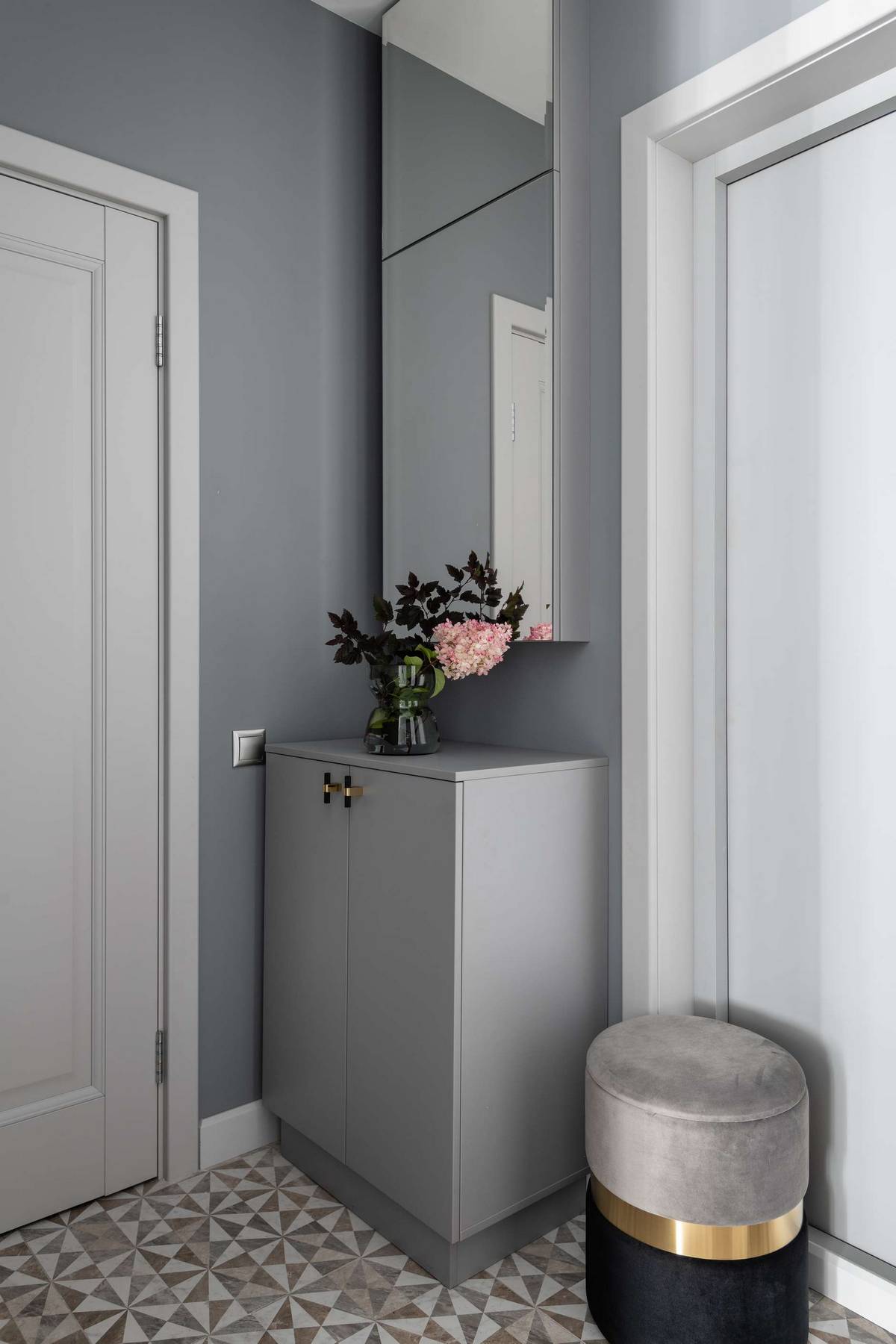 Lối vào căn hộ với chiếc tủ đựng giày màu xám đồng bộ với nền tường, lọ hoa xinh trang trí, bên trên là tủ ốp cửa gương phản chiếu không gian rộng lớn hơn.