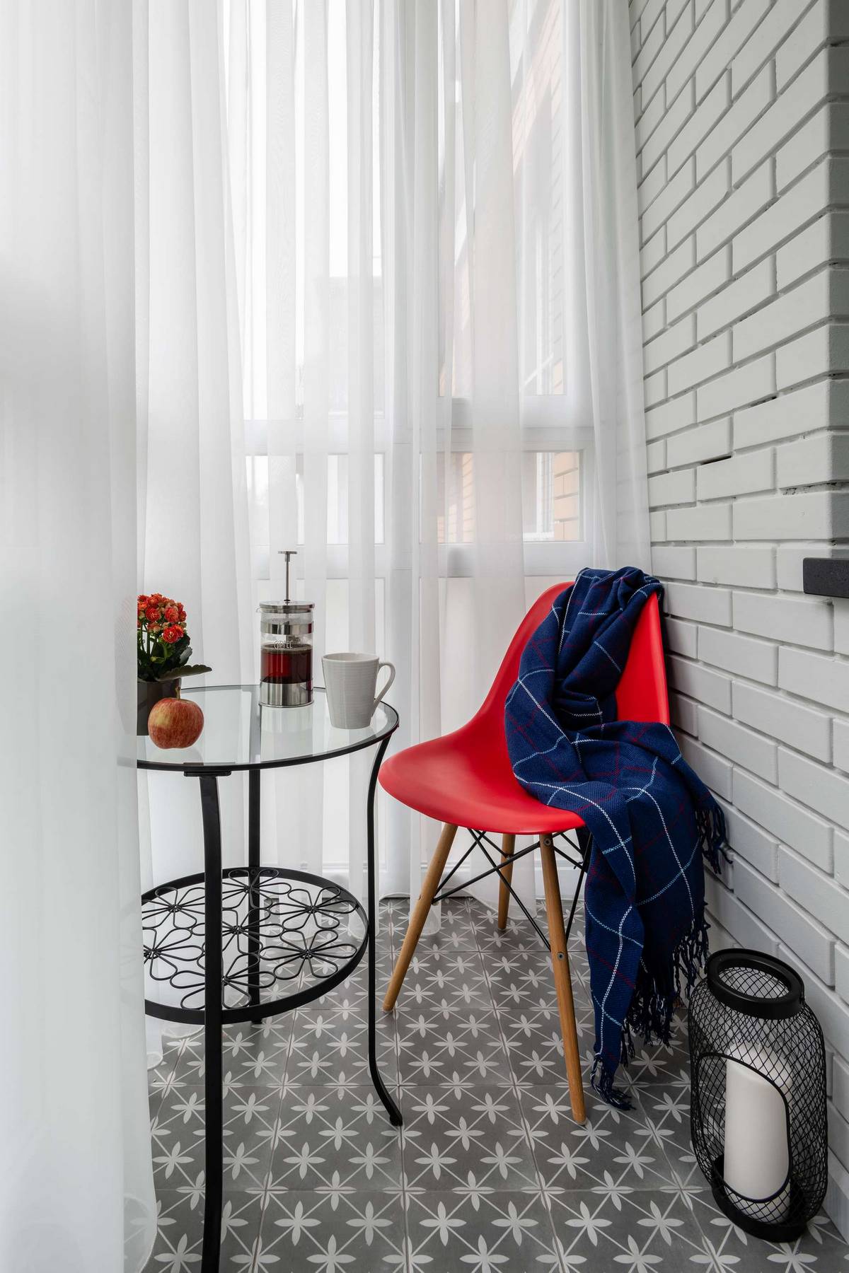 Logia trang trí theo phong cách cổ điển với tường gạch trắng, gạch bông xám, bàn nước, giỏ thắp nến kim loại sắc sảo cùng chiếc ghế đỏ nổi bật làm nơi thư giãn.