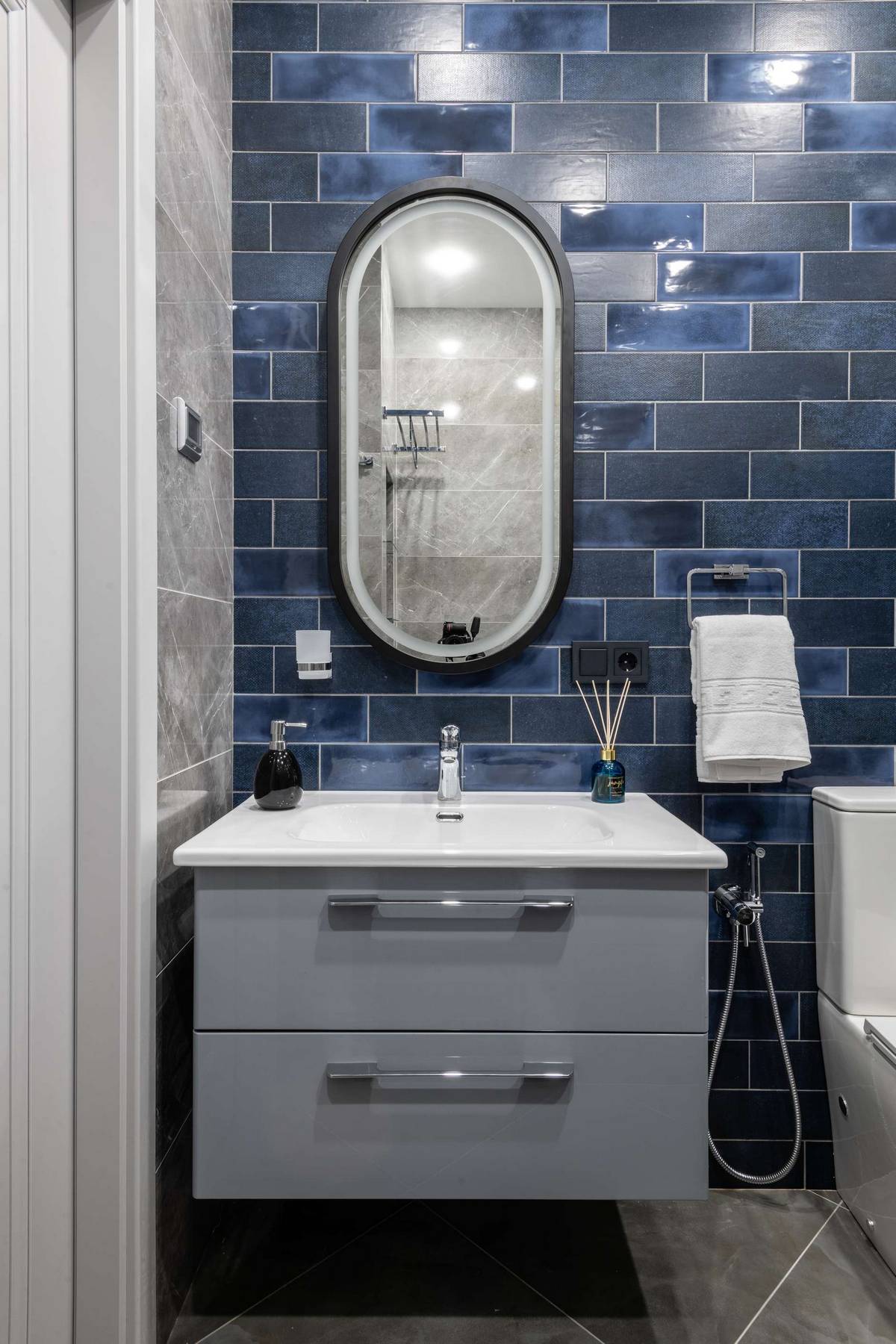 Tủ lưu trữ kết hợp bồn rửa gắn tường giúp giải phóng diện tích mặt sàn, tấm gương hình oval trên bức tường ốp gạch xanh lam đậm, nhạt đẹp mắt.