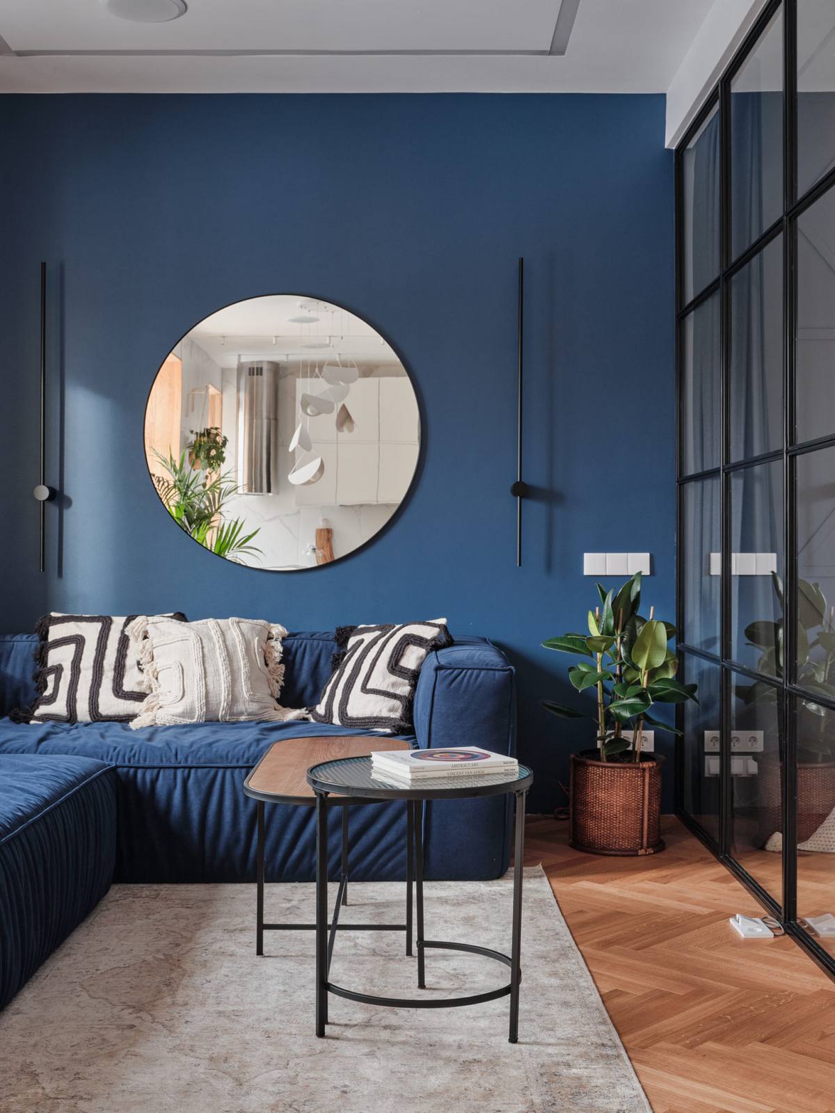Sofa góc hình chữ L màu xanh lam đồng màu với bức tường, nổi bật những chiếc gối phong cách Boho. Tấm gương tròn tạo điểm nhấn phản chiếu phòng bếp sắc trắng xinh đẹp trên nền tường sau sofa phòng khách.