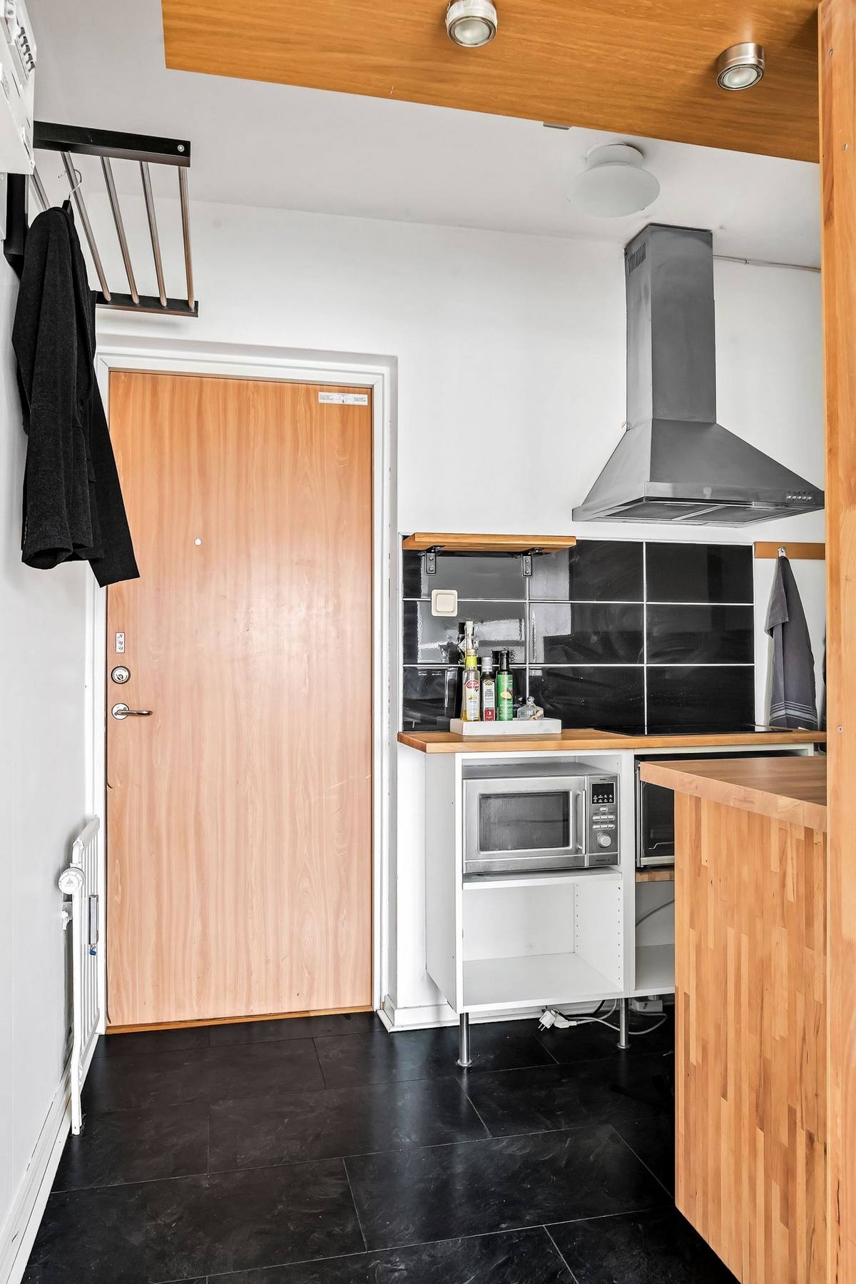 Lối vào căn hộ với cánh cửa gỗ nổi bật giữa bức tường trắng, vì diện tích rất nhỏ, chỉ hơn 20m² nên phòng bếp được bố trí ngay góc cửa ra vào, với sàn nhà màu đen tạo sự phân vùng với khu vực sinh hoạt chung phía trong.