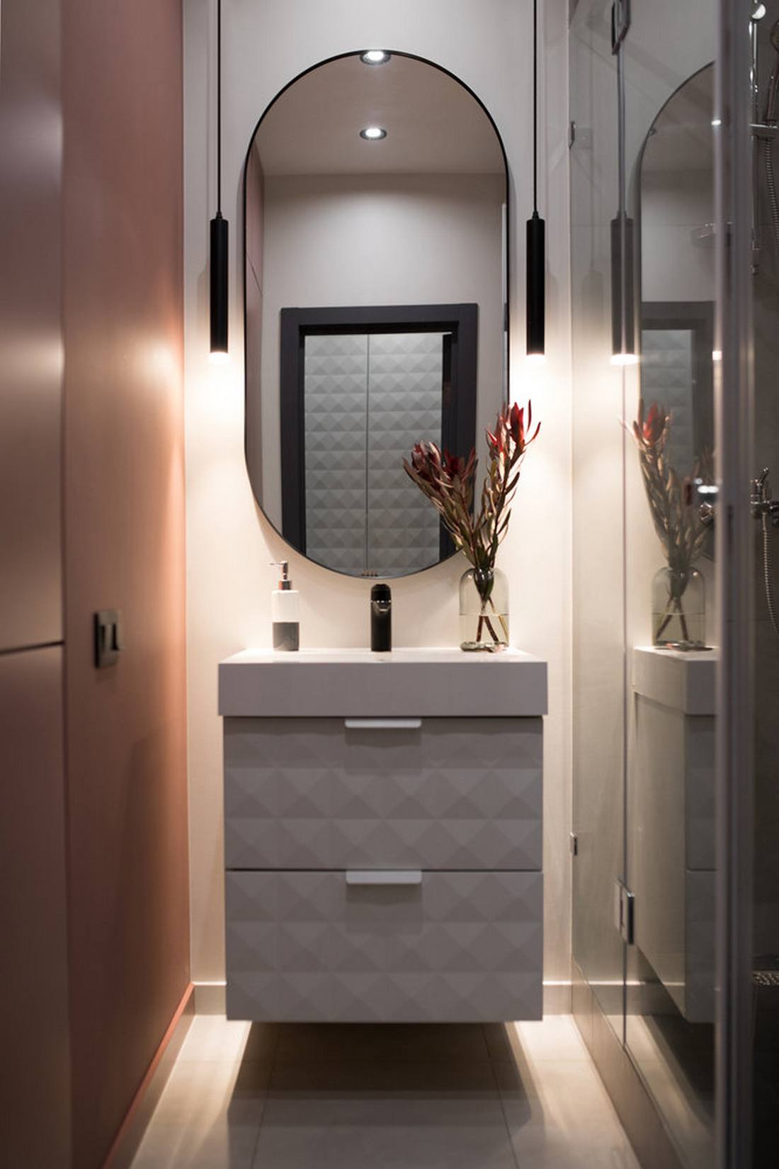 Phòng tắm nhỏ xinh đẹp với chiếc tủ lưu trữ kết hợp bồn rửa màu trắng gắn tường tiện lợi. Tấm gương hình oval trên tường phản chiếu ánh sáng từ đèn âm trần và cặp đèn thả duyên dáng, ấm áp.