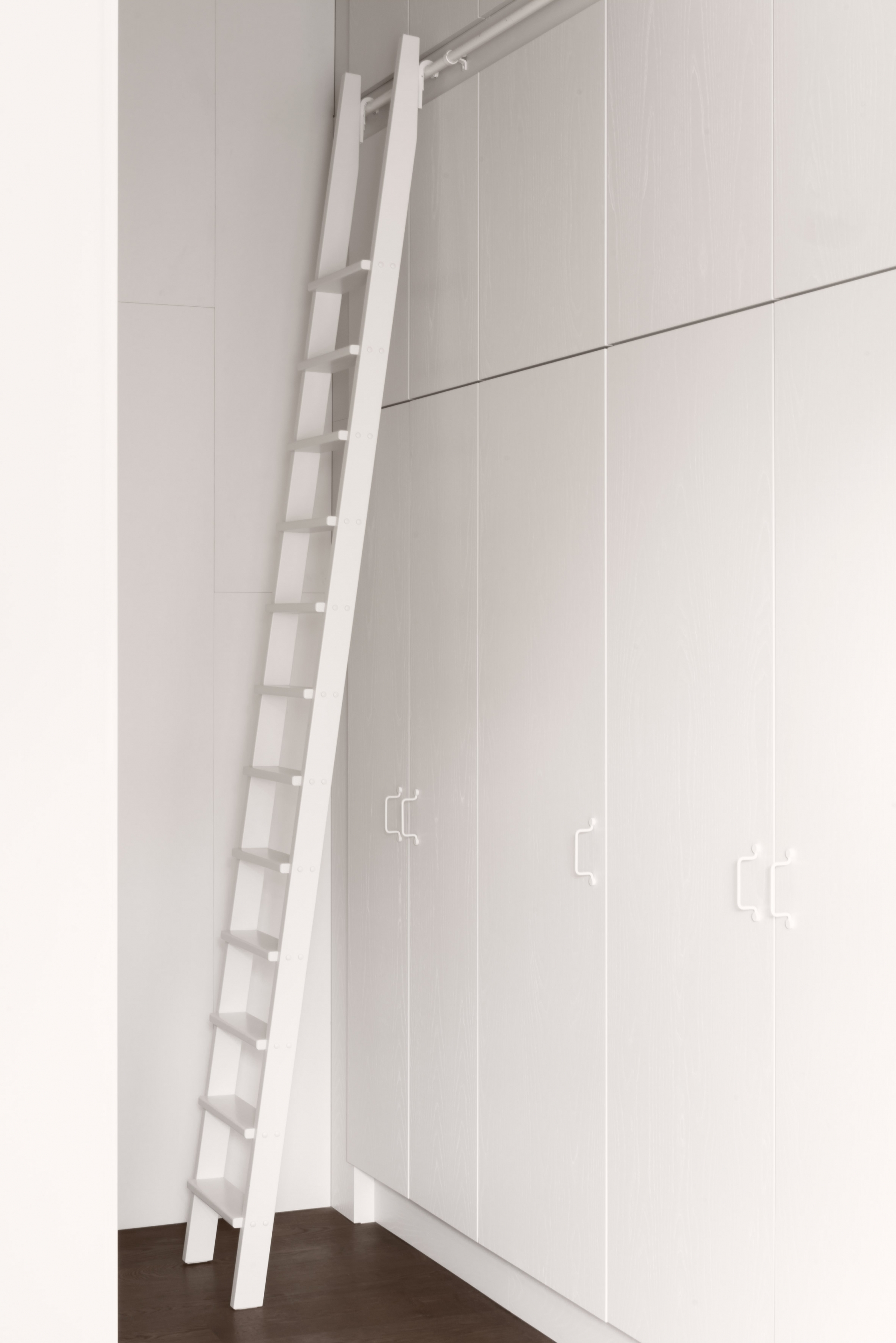 Hệ thống tủ lưu trữ với chiều cao kéo dài từ trần đến sàn nhà được lắp đặt nơi hành lang kết hợp chiếc thang gỗ đồng nhất một màu trắng tinh khôi.