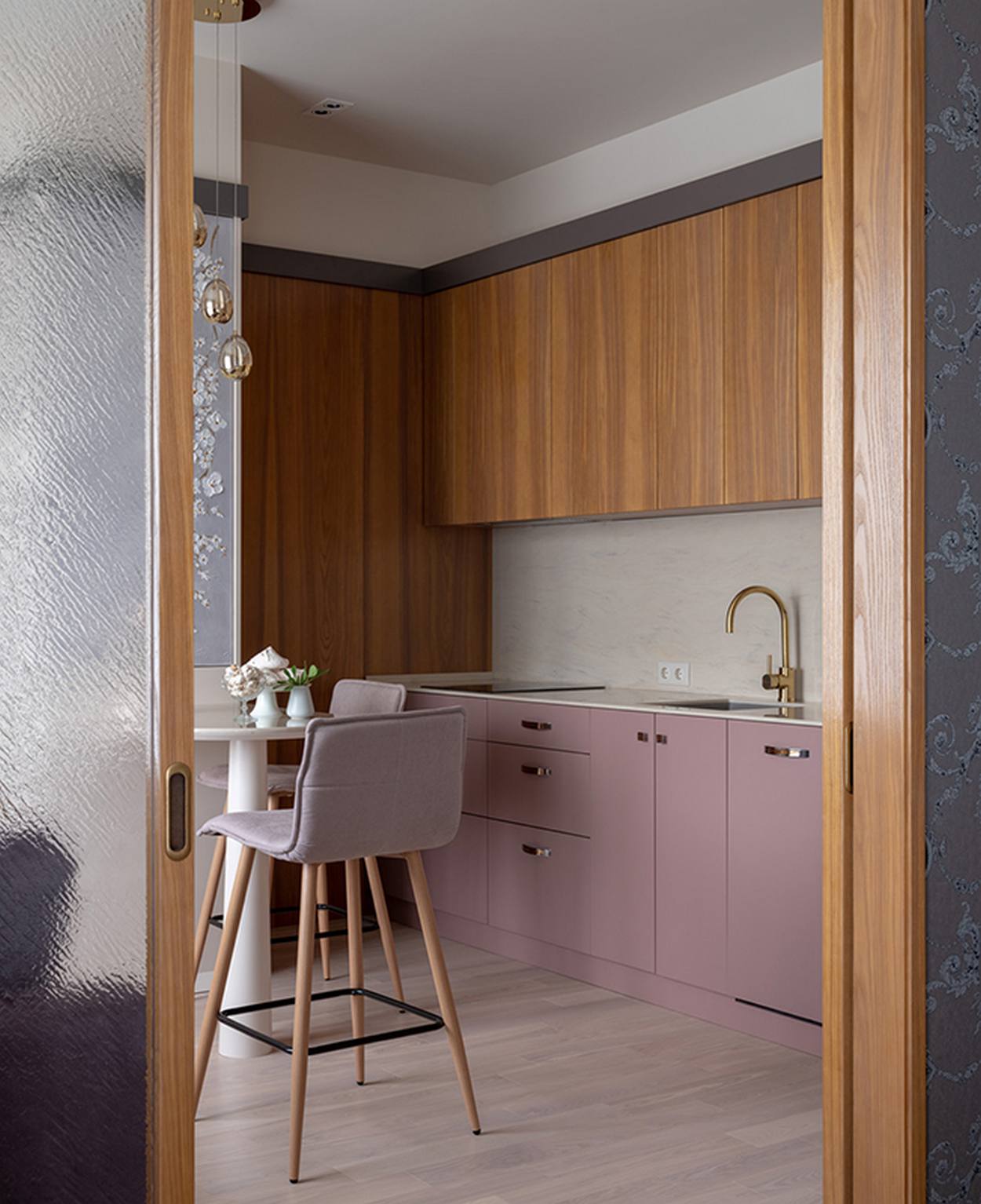 Hệ tủ bếp trên và bức tường gỗ có vân sọc dọc tạo sự đối lập nhẹ nhàng với tủ bếp bên dưới màu tím hoa cà. Backsplash ốp gạch màu trắng, cùng những chi tiết vòi rửa bằng sơn mạ vàng đẹp mắt.