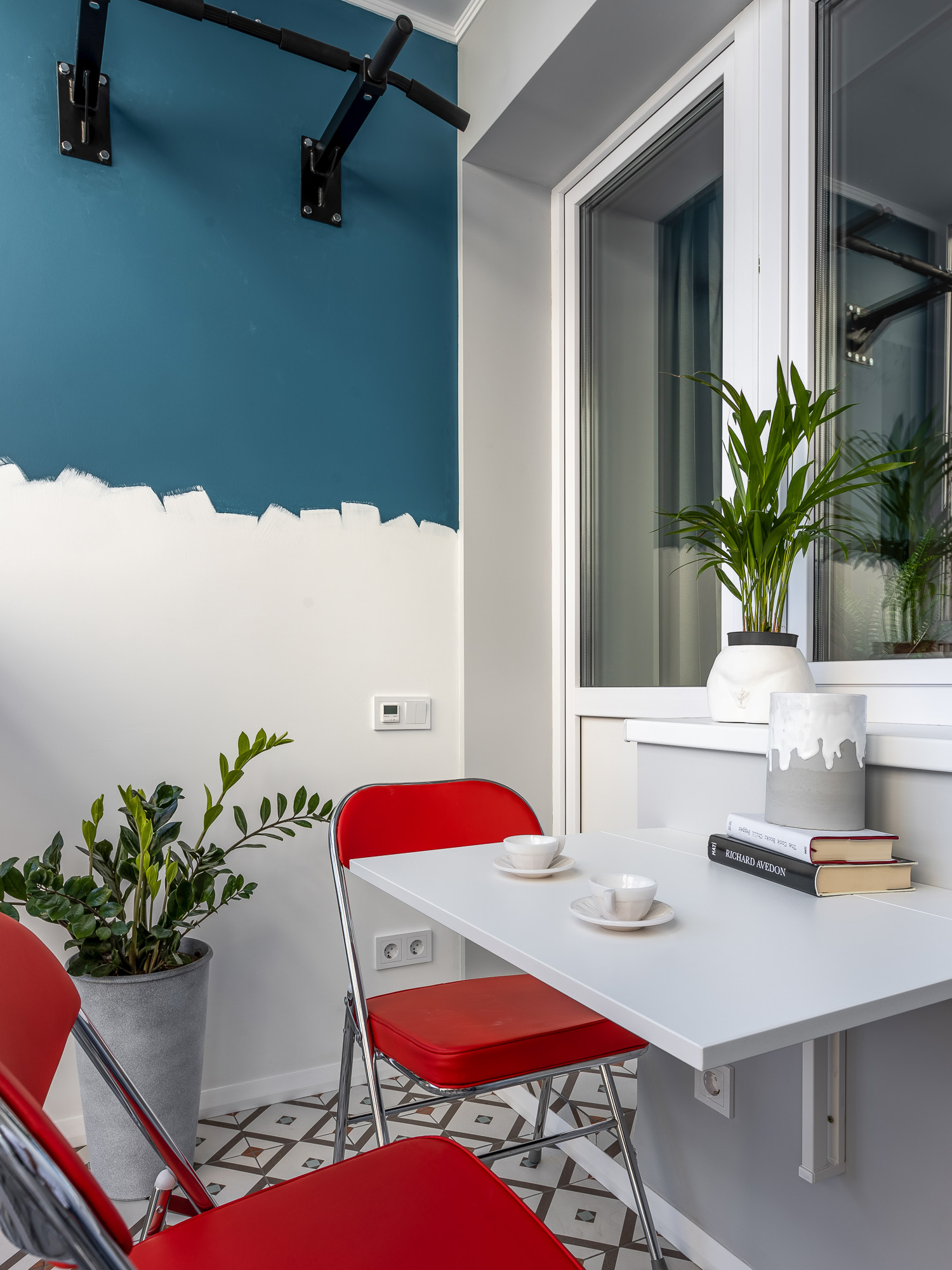 Bên ngoài là góc logia nhỏ nhắn với những chậu cây xanh trang trí cùng bức tường sử dụng cách thức sơn nửa tường trắng - xanh đầy ngẫu hứng. Những chiếc ghế màu đỏ nổi bật bên cạnh chiếc bàn có thể gấp gọn tạo nên một góc thư giãn nhẹ nhàng nhưng cá tính.