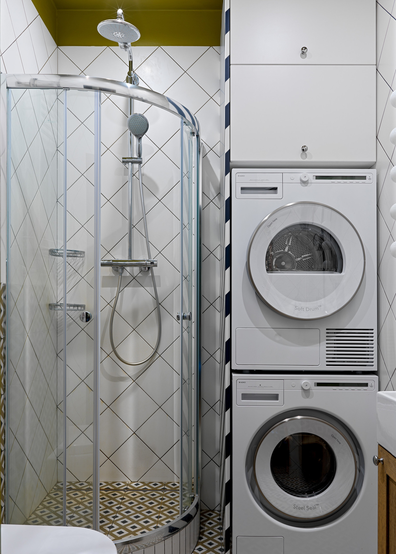 Vì diện tích phòng tắm không quá rộng rãi nên các thiết bị như máy giặt, máy sấy được xếp chồng lên nhau, phía trên cùng là tủ lưu trữ. Buồng tắm thiết kế trong góc nhỏ, phân vùng bởi cửa kính trượt trong suốt theo đường cong mềm mại.