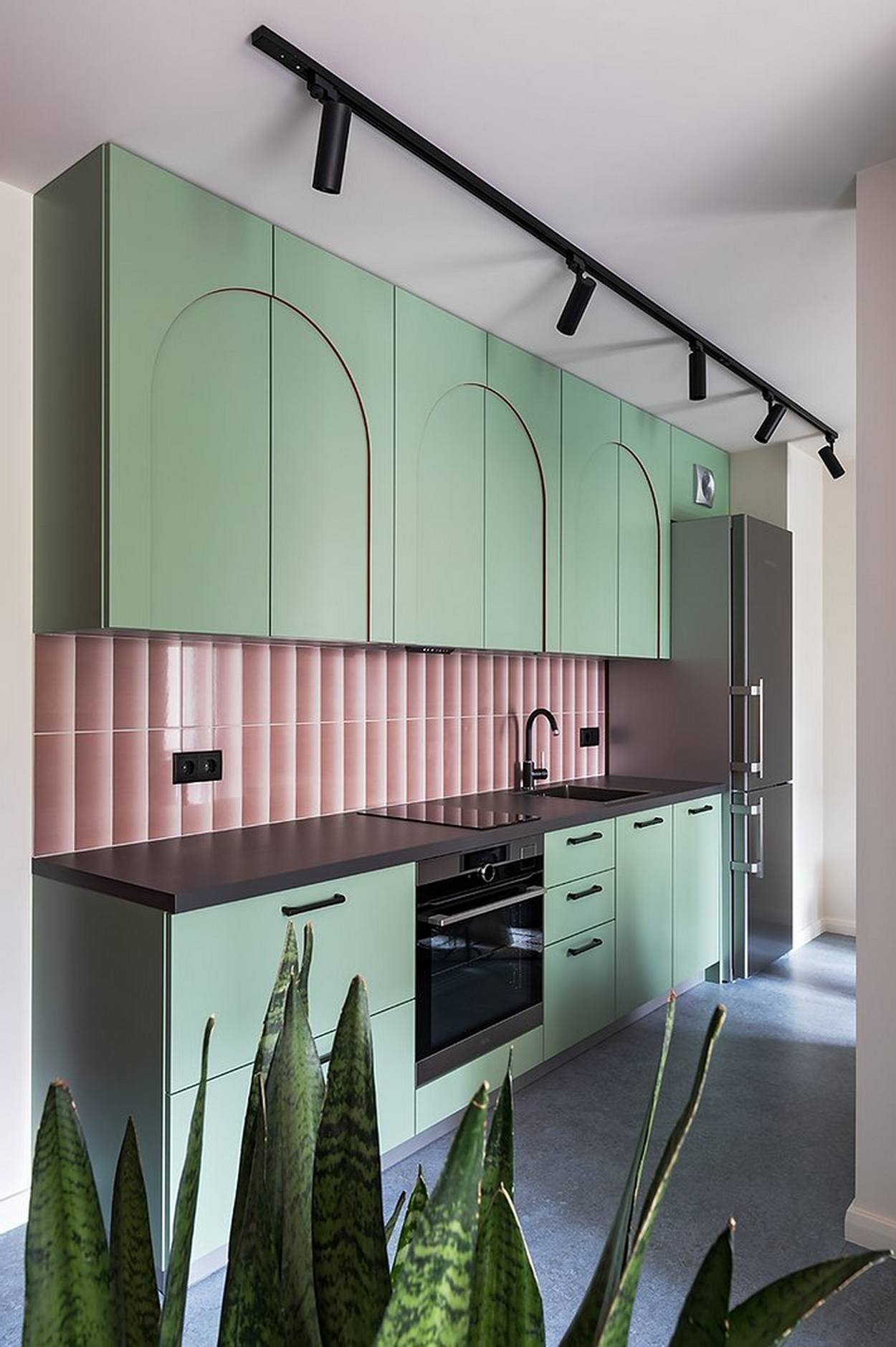 Phòng bếp thiết kế kiểu chữ I, phù hợp với cấu trúc dài hẹp, tạo lối đi thông thoáng đồng thời đảm bảo tiện nghi cho không gian nấu nướng. Khu vực backsplash ốp gạch hồng sáng bóng làm điểm nhấn giữa hệ tủ bếp màu xanh bạc hà dịu mát.