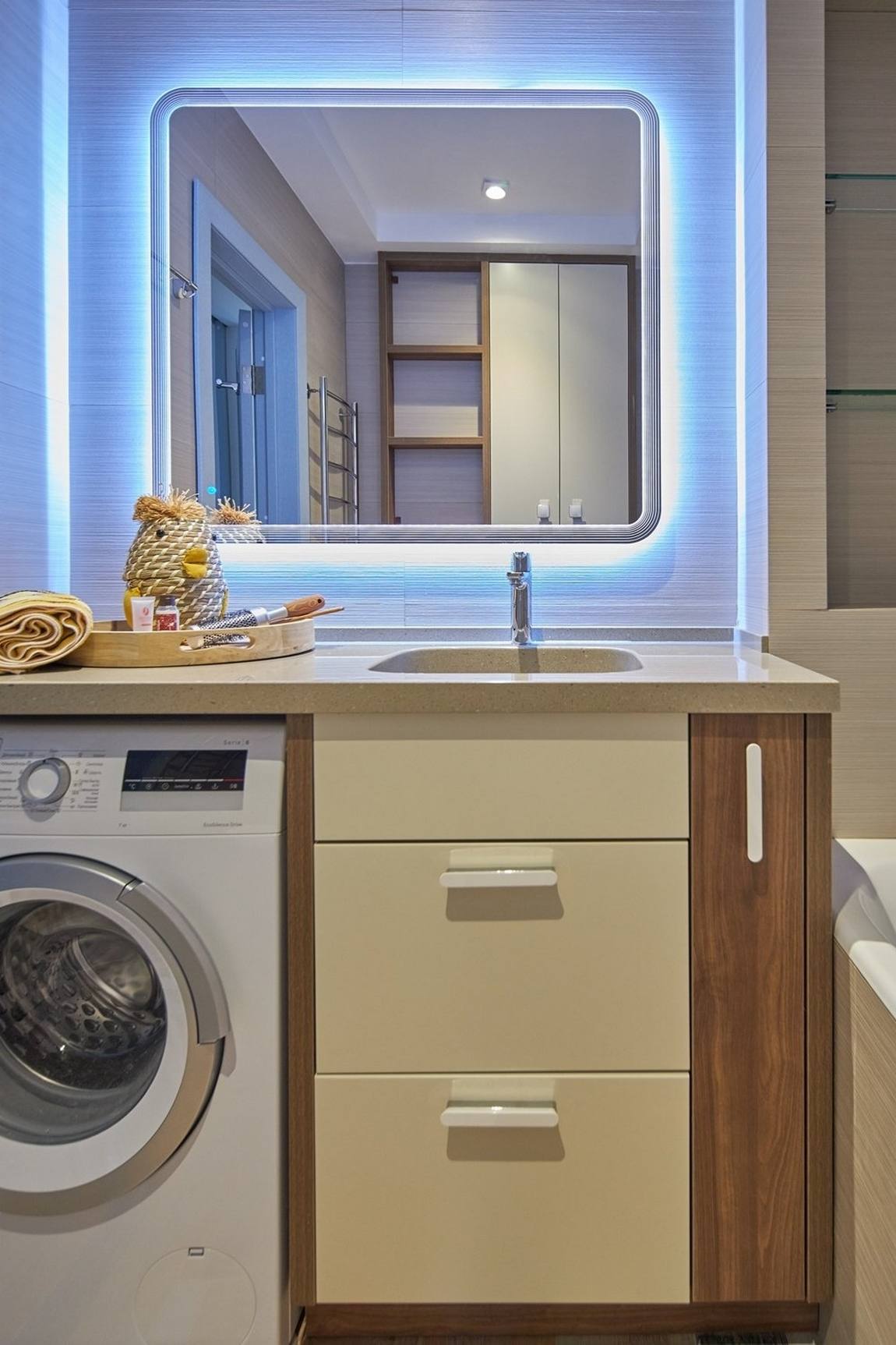 Phòng tắm với máy giặt bố trí gọn gàng bên dưới tủ lưu trữ kết hợp bồn rửa. Trên tường là tấm gương hình vuông với các góc cạnh bo tròn, xung quanh viền hệ thống đèn LED bắt mắt.