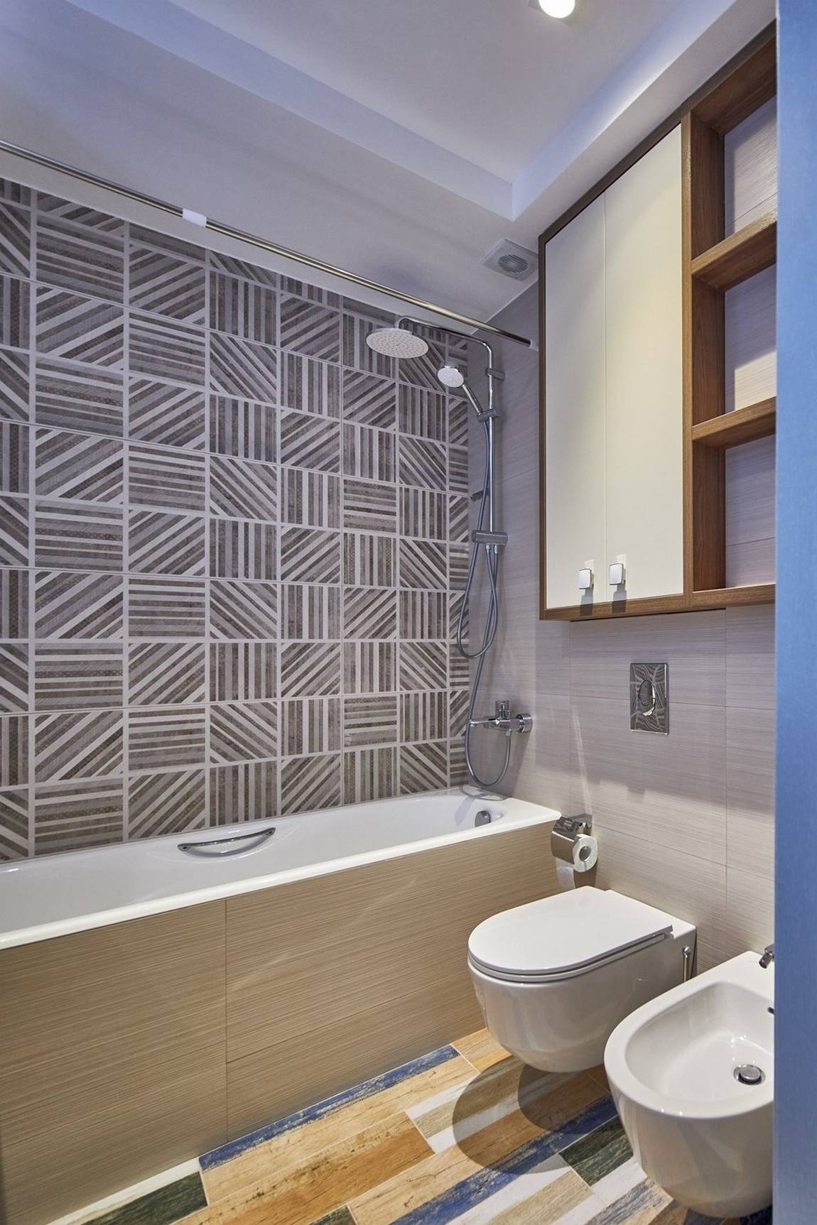 Sàn phòng tắm lát gạch giả gỗ nhiều màu sắc nổi bật, trên tường tích hợp tủ kệ lưu trữ và những viên gạch họa tiết hình học xen kẽ trên tường bồn tắm. Toilet gắn tường cũng giúp cho không gian nhỏ dễ vệ sinh chùi rửa hơn.