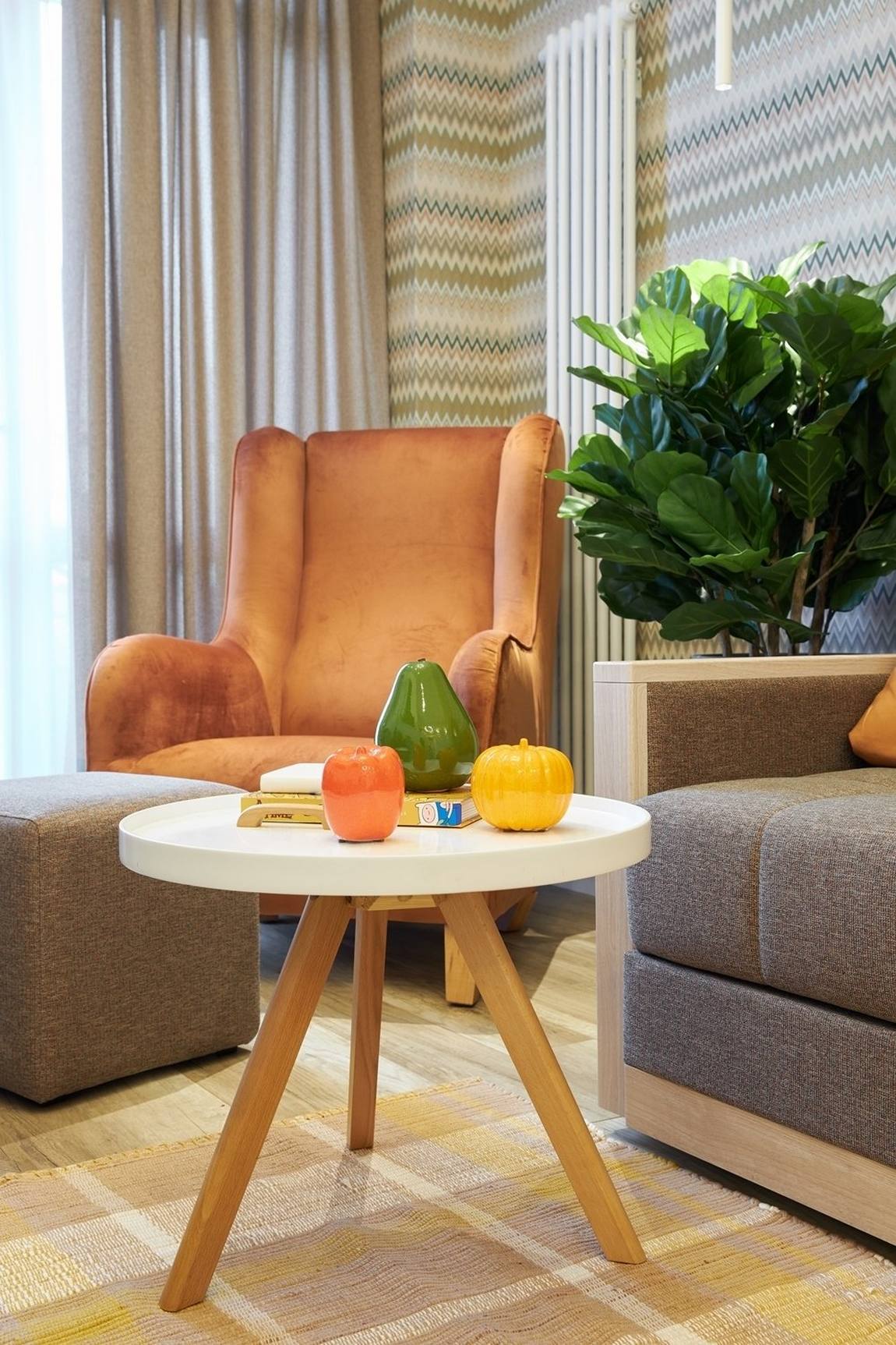 Bàn nước thiết kế đơn giản với mặt bàn tròn màu trắng, chân ghế gỗ đặt trên tấm thảm sáng màu. Chiếc ghế bành màu cam gạch đặt ở góc cửa sổ tạo nên góc thư giãn nhẹ nhàng. Chậu cây cảnh tươi xanh cũng giúp cho căn phòng thêm sinh động.