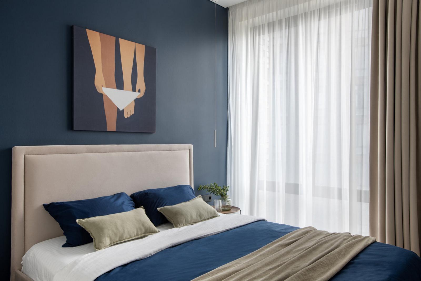 Phòng ngủ cũng sở hữu cửa sổ đầy nắng ấm với 2 lớp rèm che mỏng, dày để cách âm và tạo sự riêng tư khi cần. Đầu giường vững chãi với gam màu be thanh lịch. Sắc xanh lam bao phủ bức tường, ga gối và mềm để tạo điểm nhấn.
