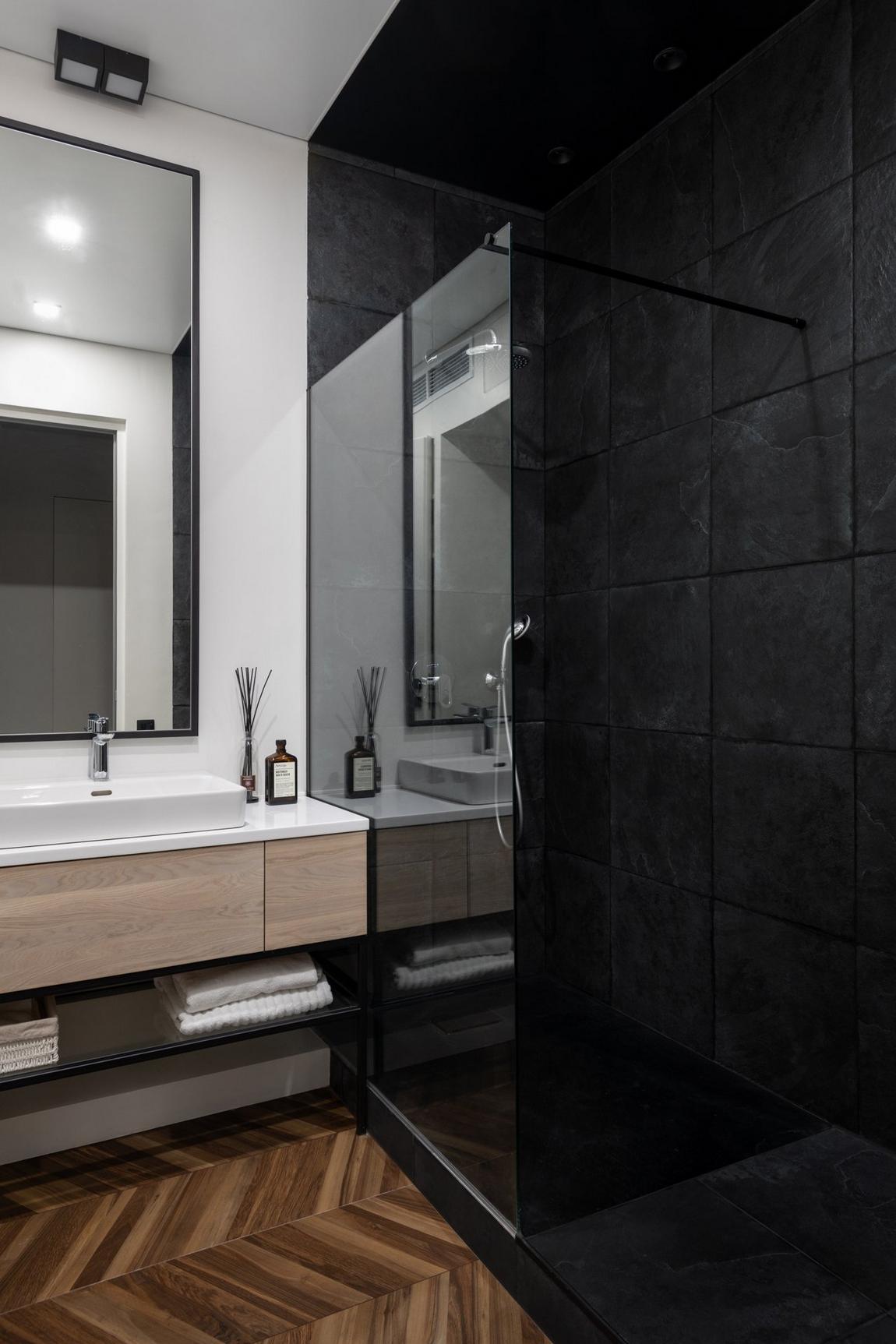 Phòng tắm và nhà vệ sinh được phân vùng bởi gạch lát sàn màu gỗ họa tiết xương cá và màu đen bí ẩn, cách nhau bởi cửa kính trong suốt. Tấm gương lớn trên tường bồn rửa góp phần 'nhân đôi' diện tích và ánh sáng cho căn phòng không cửa sổ.