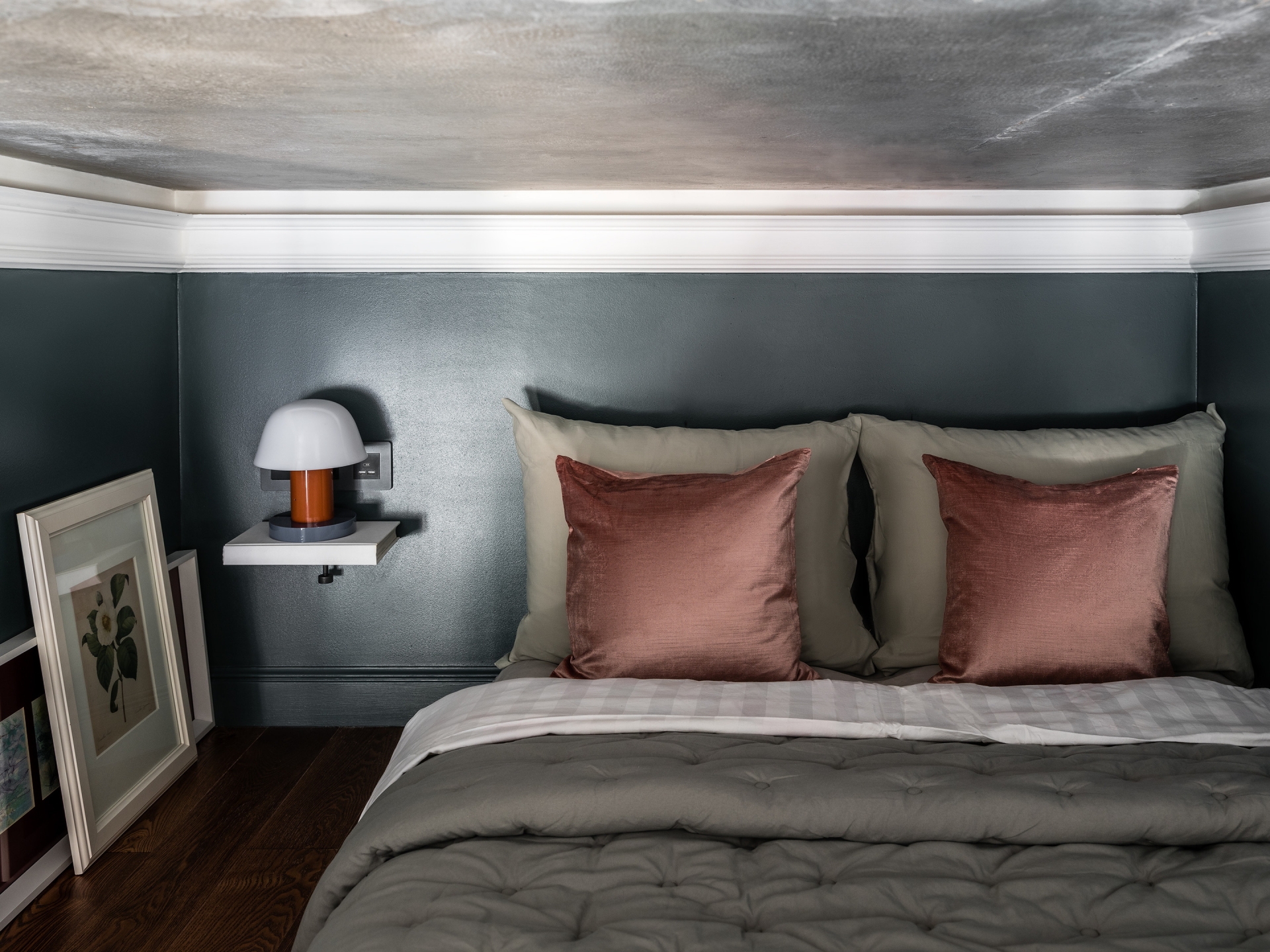 Phòng ngủ đơn giản với sàn gỗ tối màu, tường sơn xám, gối màu hồng đỗ nổi bật. Táp đầu giường nhỏ, gắn trên tường để tiết kiệm diện tích sàn, vài bức tranh hoa lá trang trí cho phòng ngủ thêm xinh.