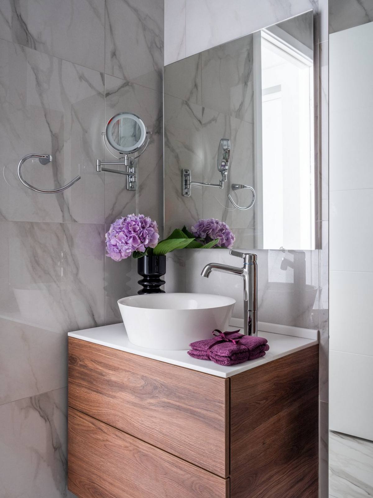 Nhà vệ sinh và buồng tắm được phân vùng bởi cửa kính trong suốt. Trên bồn rửa kết hợp tủ gỗ là chiếc lọ màu đen với cành hoa tím trang trí tương đồng với lối vào căn hộ.