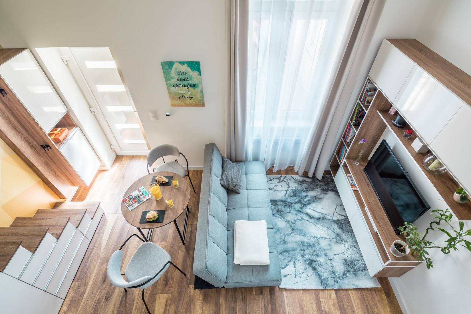 Hình ảnh phòng khách được chụp từ trên cao với ghế sofa màu xám êm ái, phân vùng với các khu vực còn lại bằng một tấm thảm trải sàn ấn tượng. Rèm che cửa sổ được tích hợp 2 lớp mỏng và dày để cản nắng và tạo sự riêng tư.