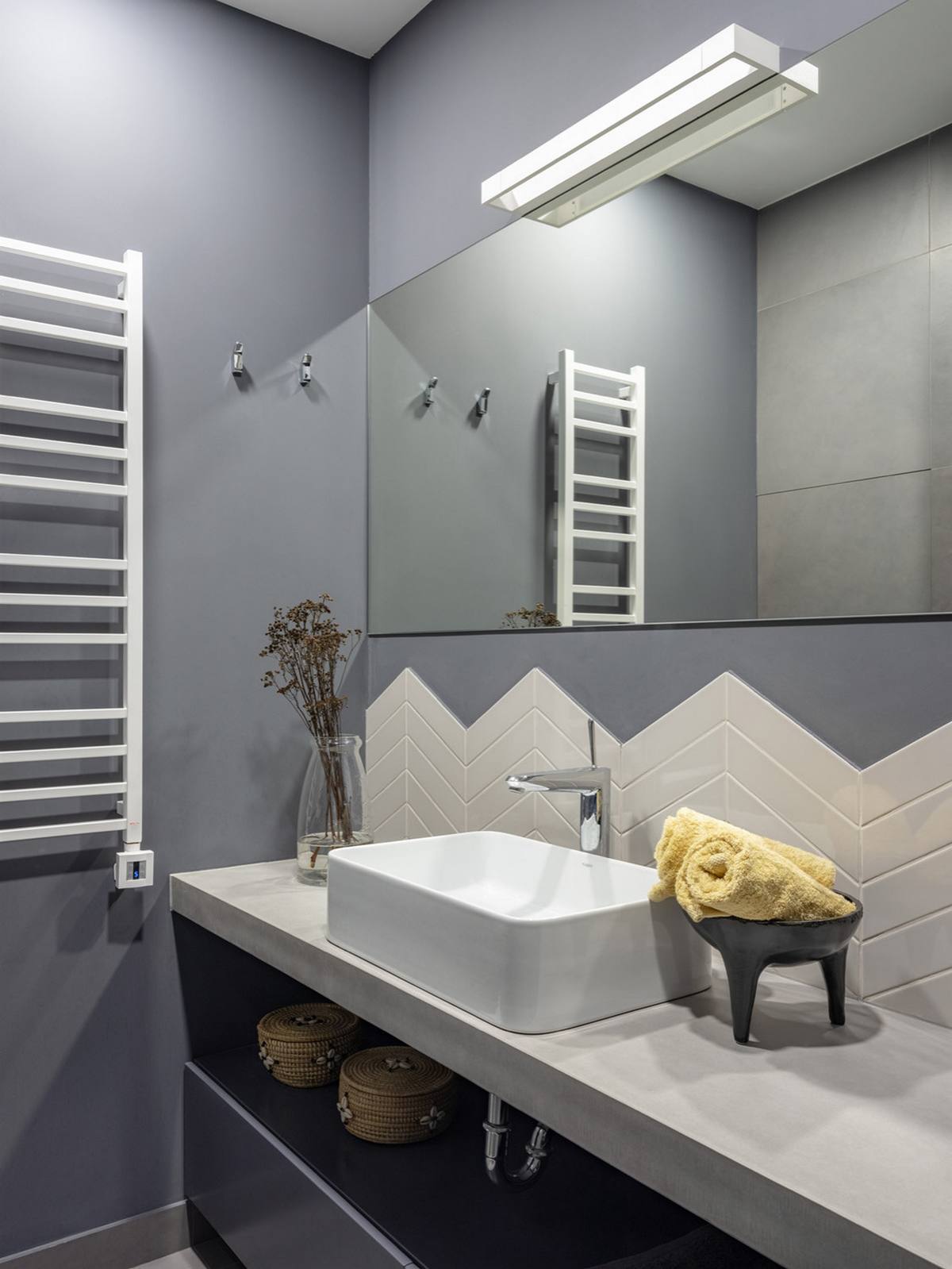 Phòng tắm kết hợp sơn tường xám nhạt chủ đạo với gạch ốp họa tiết xương cá màu trắng sáng bóng tạo điểm nhấn thu hút. Bức tường trên bồn rửa ốp gương cỡ lớn có thiết kế đơn giản, không viền khung, nhằm 'nhân đôi' không gian bằng thị giác.