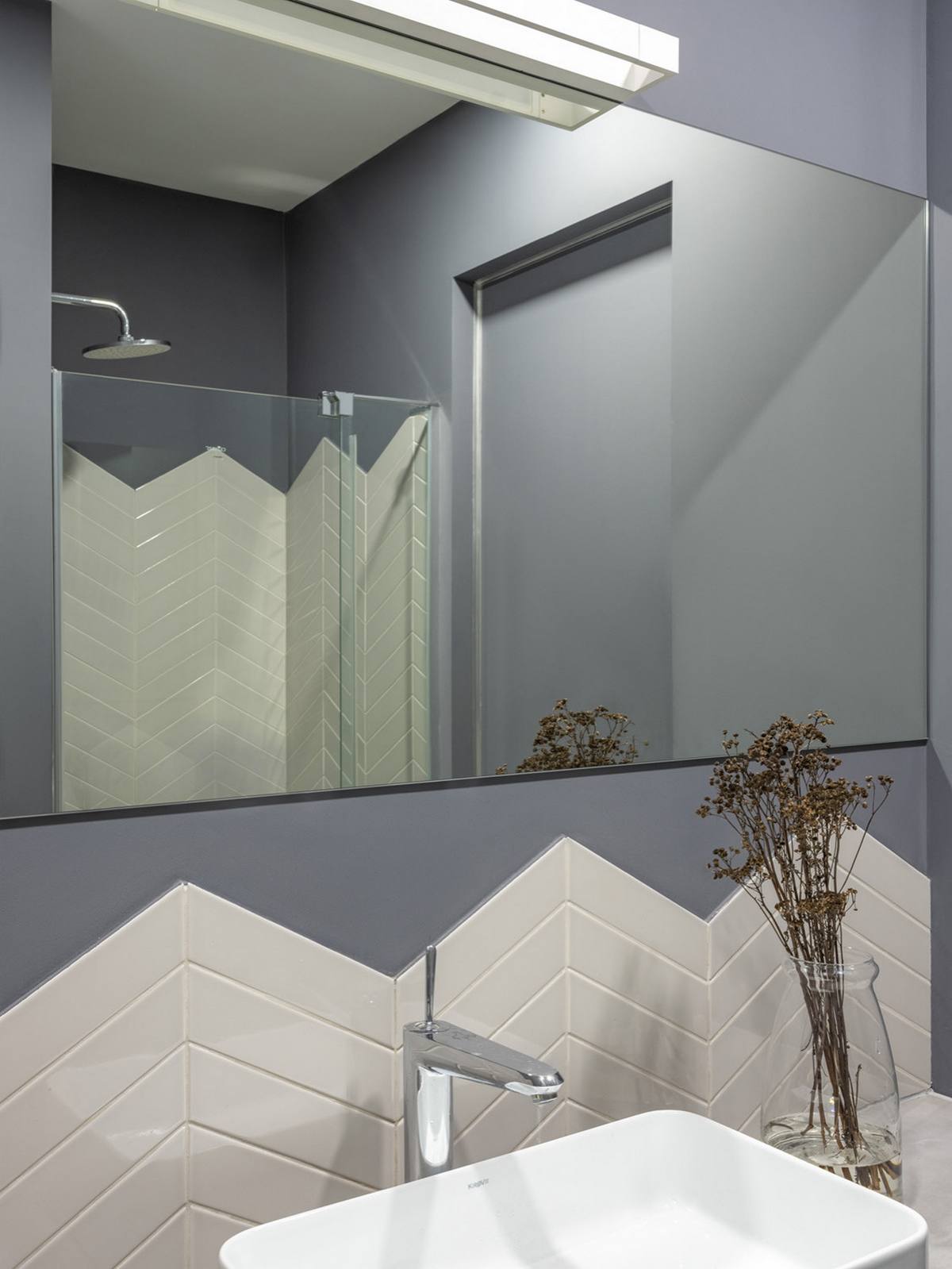 Thông qua hình ảnh phản chiếu từ tấm gương, chúng ta có thể thấy buồng tắm và nhà vệ sinh được phân vùng bởi cửa kính trong suốt. Bức tường cũng được ốp gạch họa tiết xương cá sáng bóng tạo sự liên kết hài hòa với khu vực bồn rửa tay.