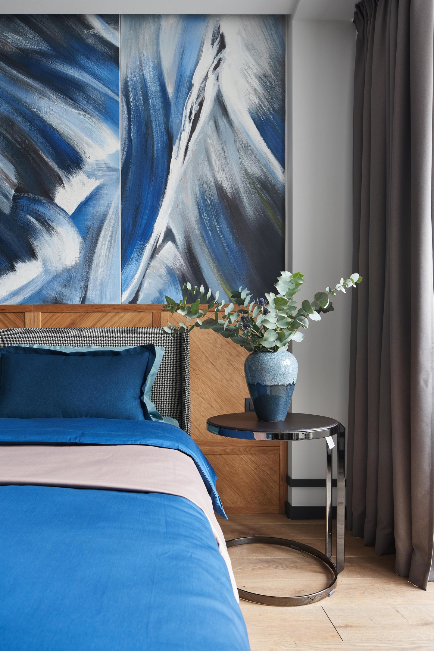 Khu vực đầu giường với tấm gỗ vững chãi làm điểm tựa đồng thời cũng tăng thêm sắc màu cho không gian. Táp đầu giường bằng kim loại sáng bóng, bên trên là lọ hoa bằng gốm sứ có gam màu xanh lam đậm - nhạt hài hòa và tinh tế.